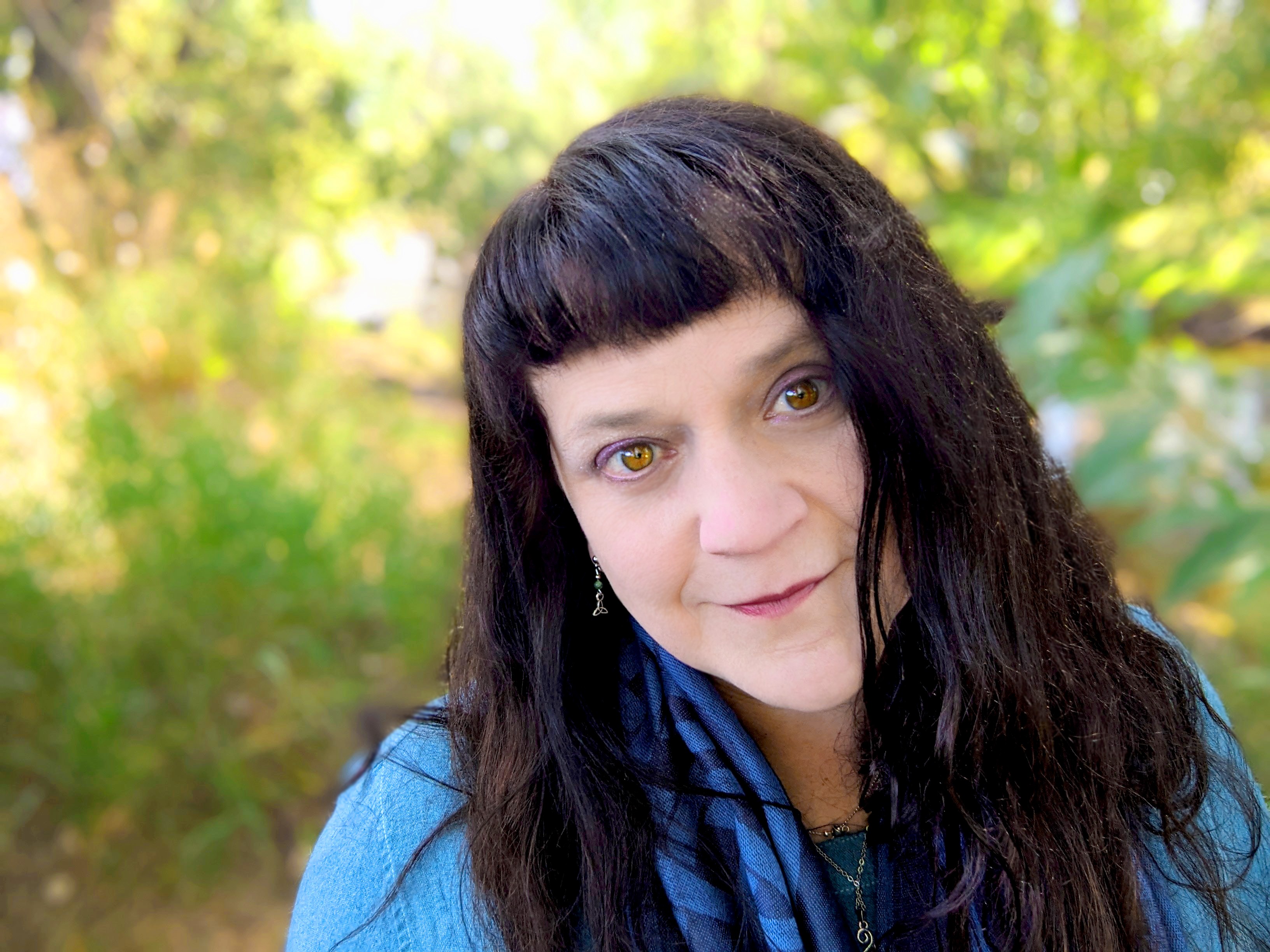 Woman with dark wavy hair and bangs outdoors, wearing a blue jacket, smiling at the camera.
