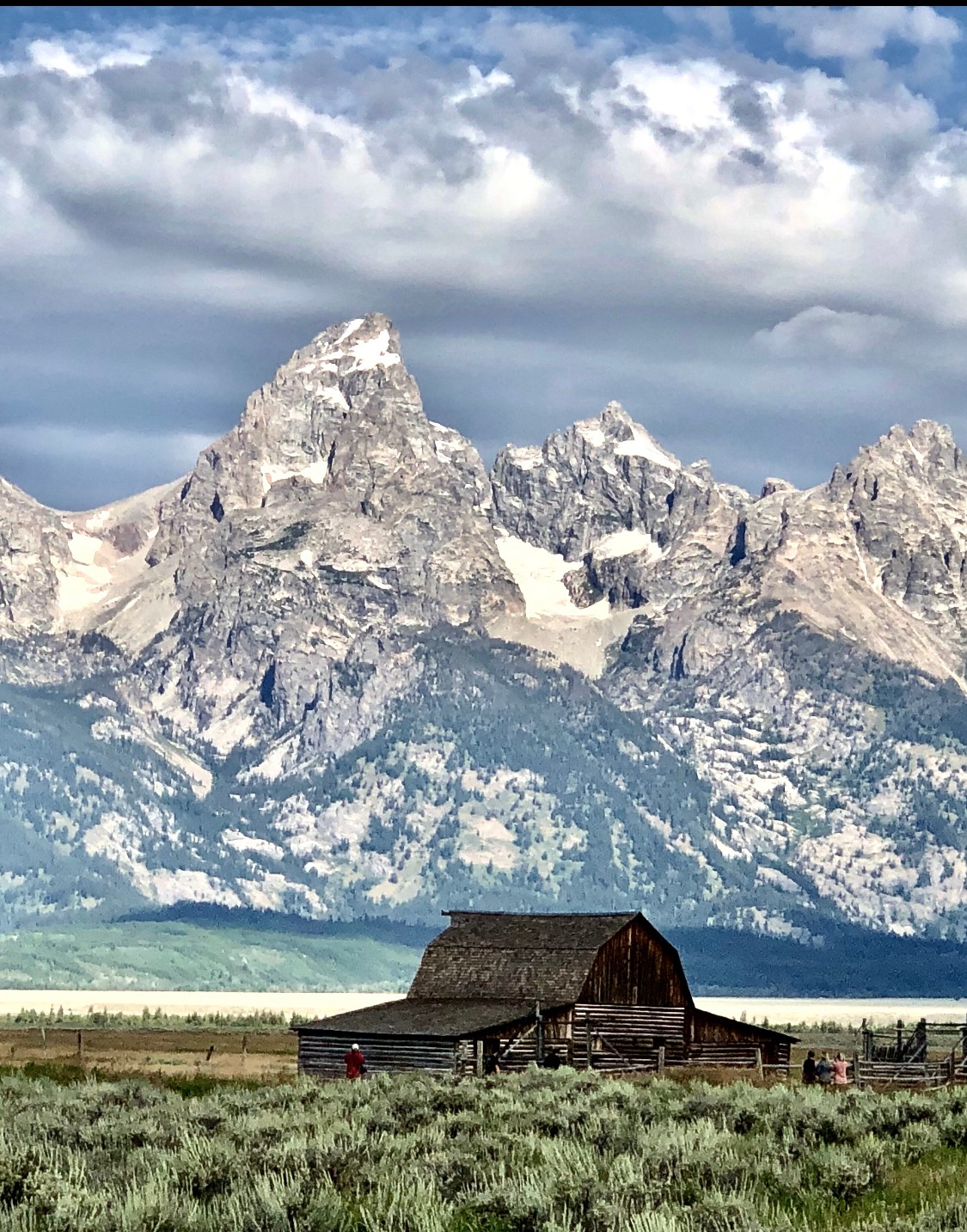 Snow-capped mountain range with a rustic barn in the foreground, cloudy sky, and grassy field.