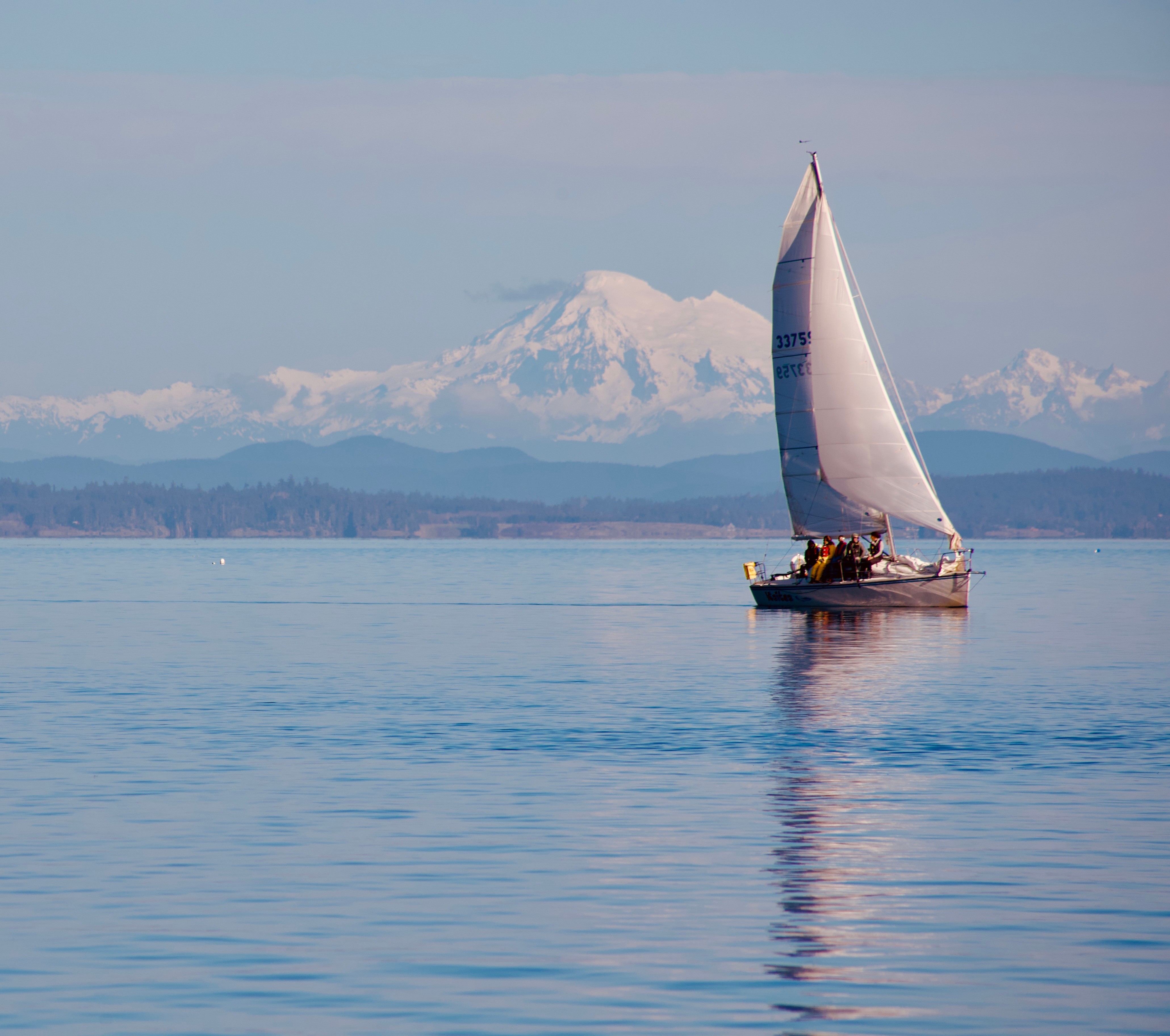 Sailboat on calm water with snow-capped mountain in background, clear sky, and distant shoreline.