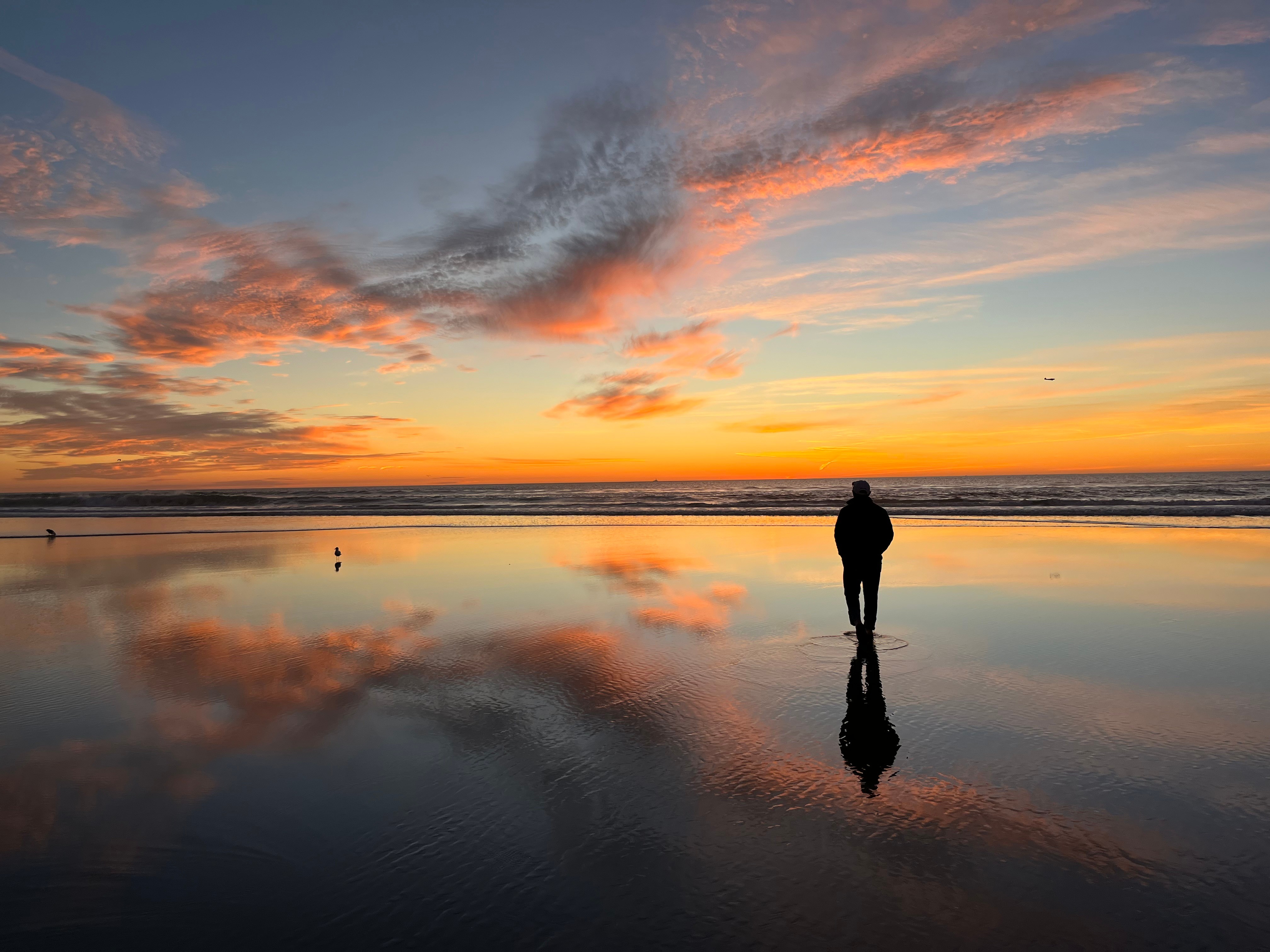 Person walking on a beach at sunset with colorful sky and reflection on wet sand.