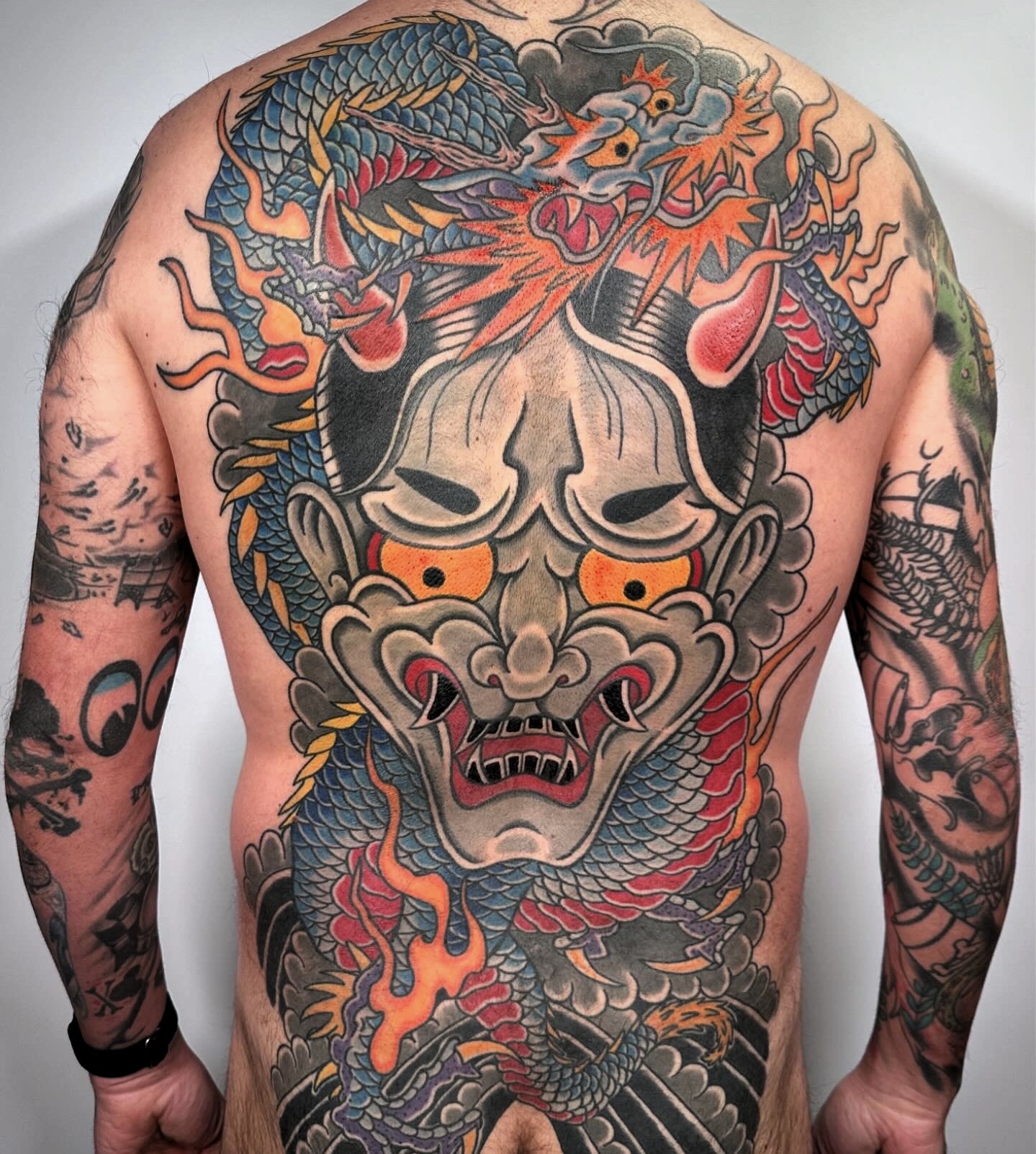 Large colorful tattoo of a fierce demon face with flames, a dragon, and clouds covering the person's back and arms.