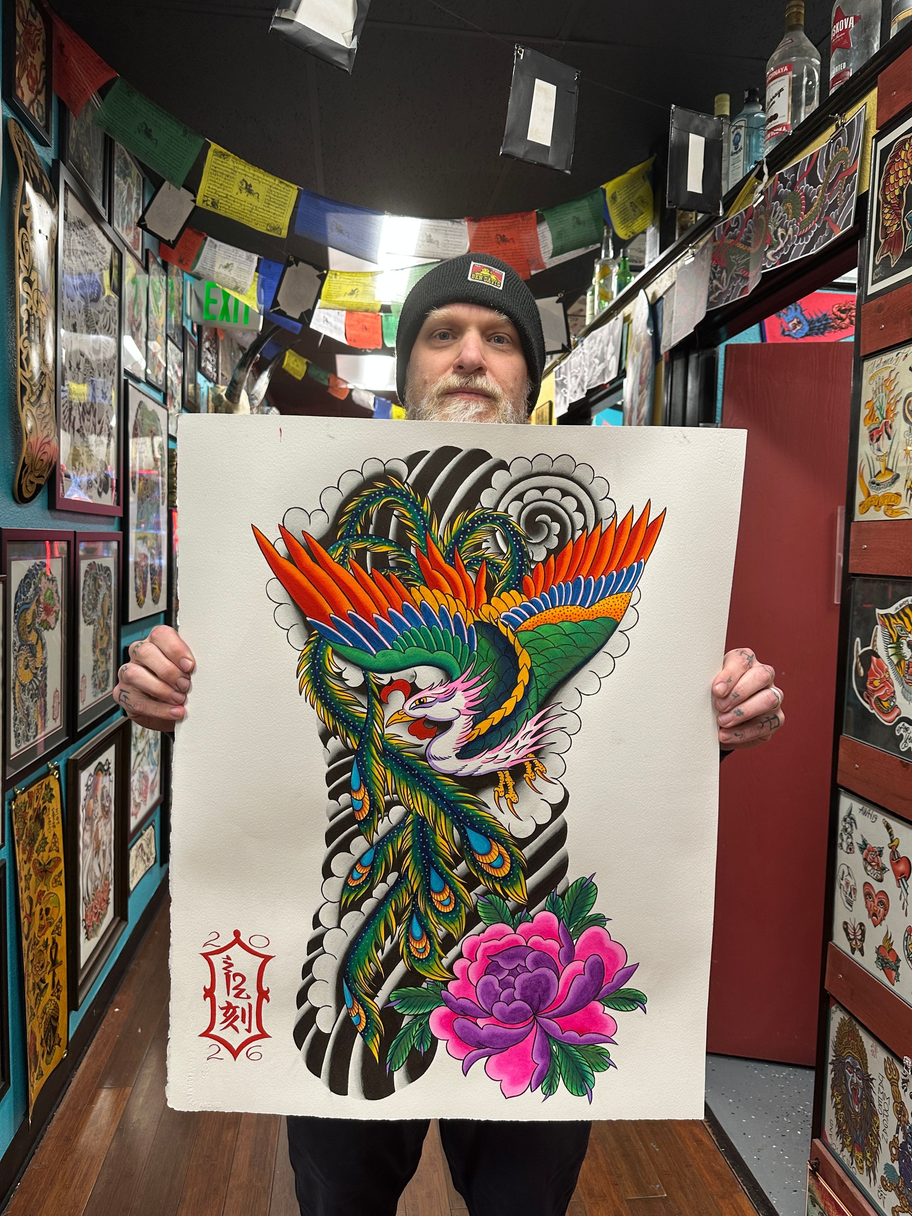 Person holding a colorful tattoo-style dragon and bird illustration with a pink flower, in a store with framed artwork.