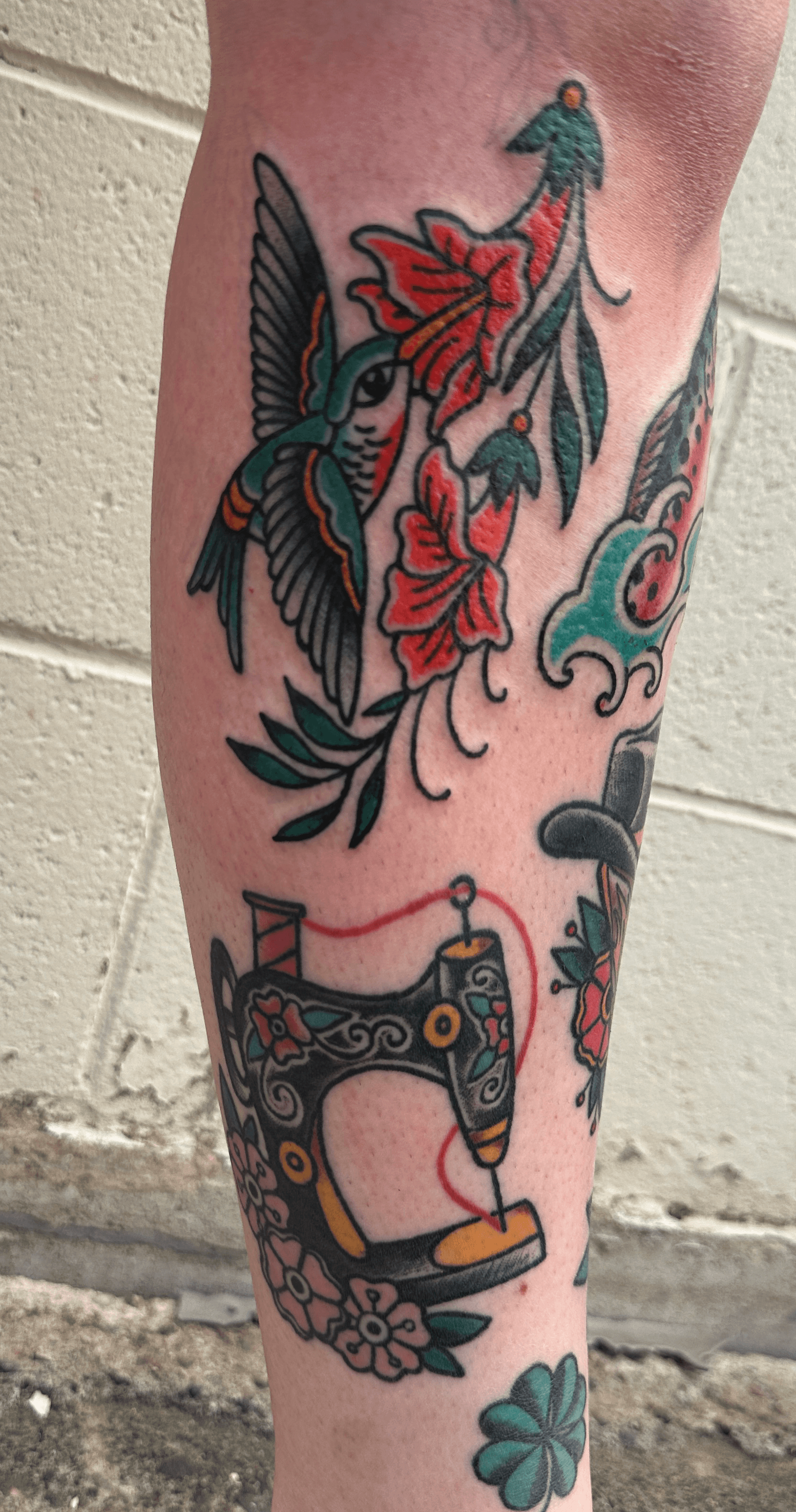 Tattoo of a hummingbird, red flowers, and a vintage sewing machine on a person's leg.