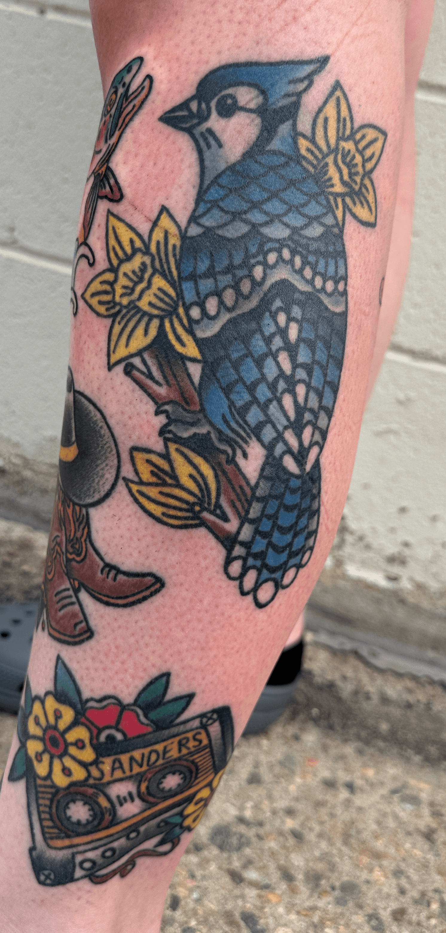 Colorful tattoo of a bird with blue and black feathers surrounded by yellow flowers on skin.