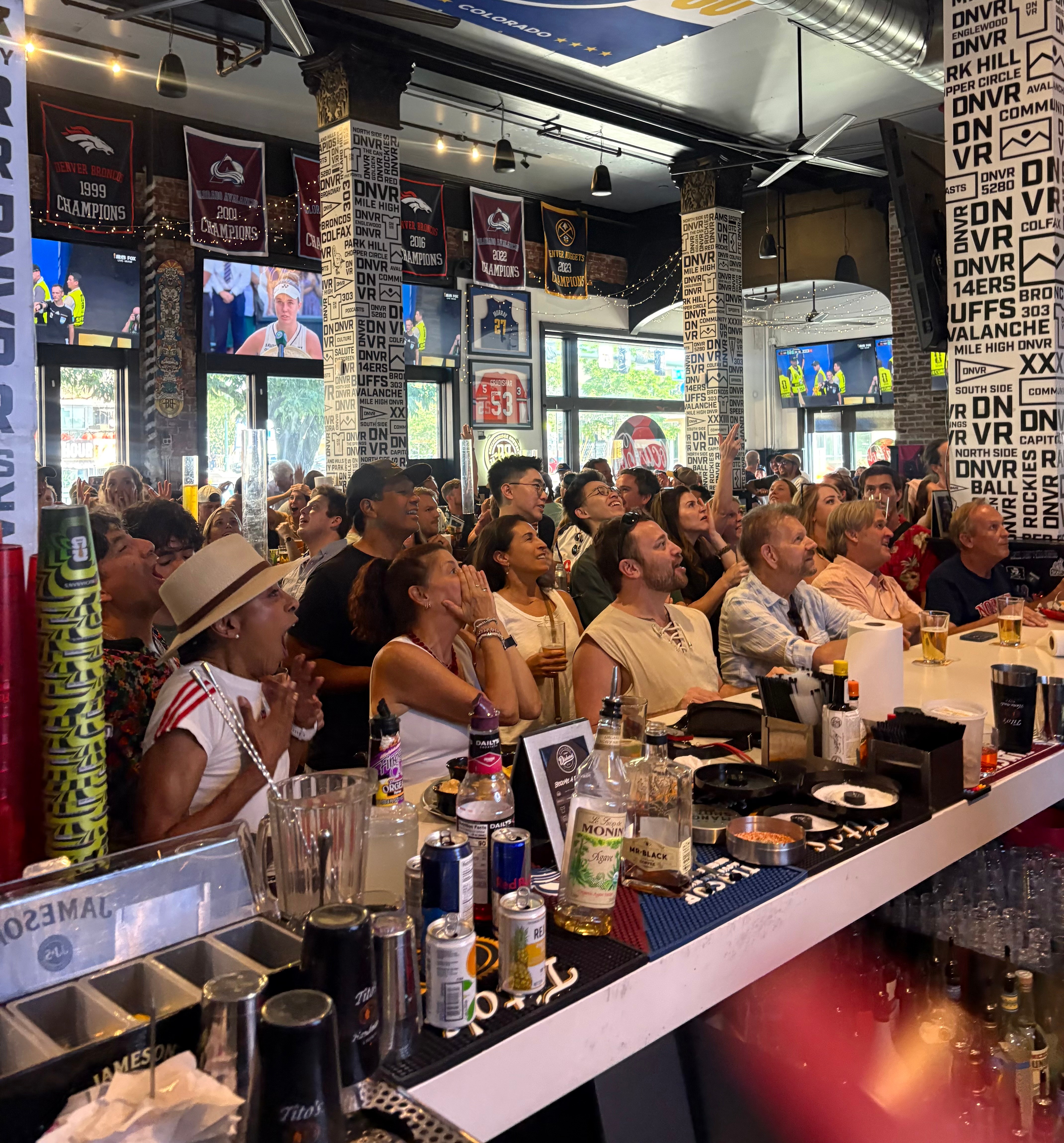 Crowd of people watching a screen in a bar or restaurant, with tables, drinks, and posters on the walls.