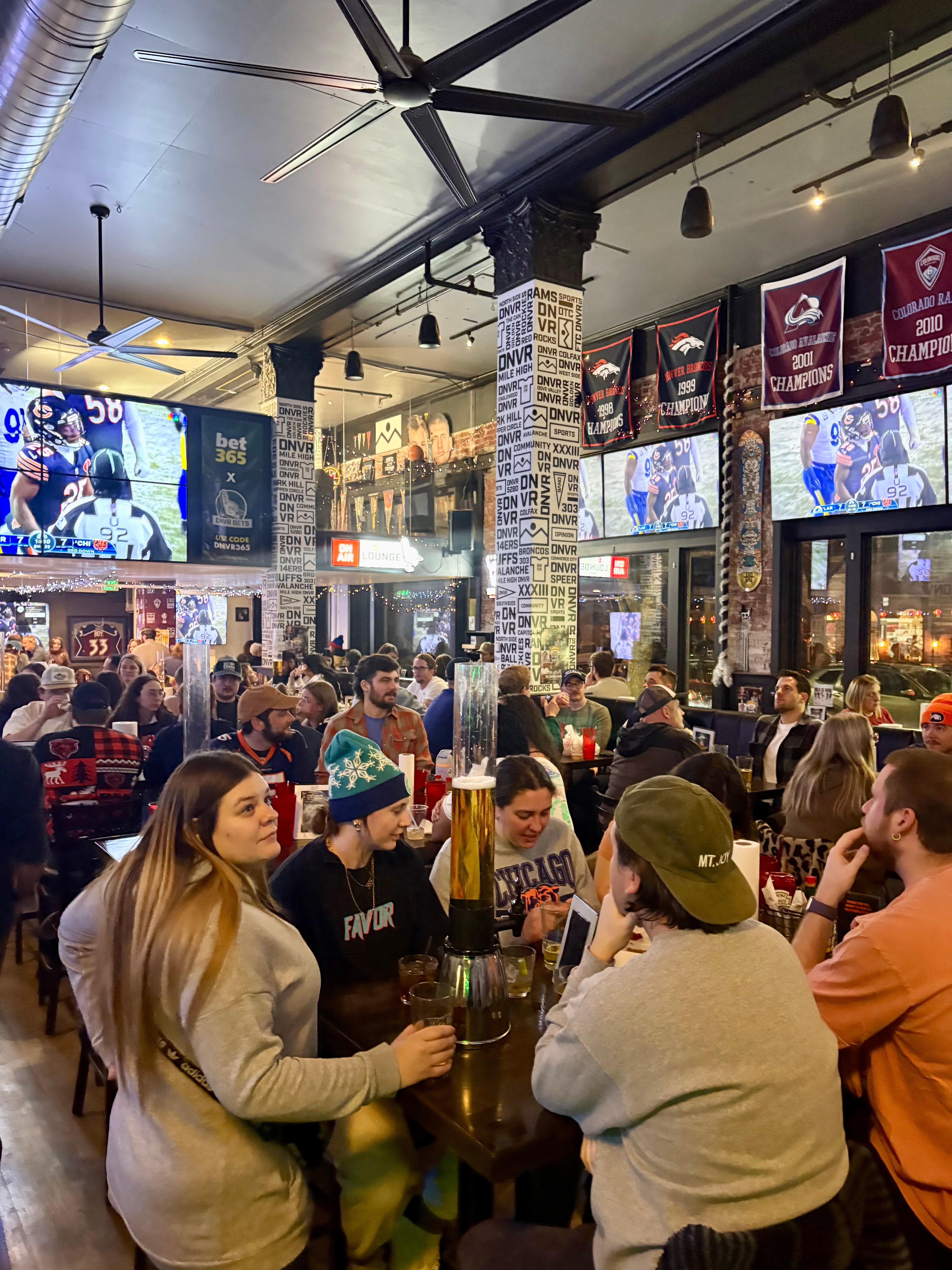 Crowded sports bar with people watching TV screens, sitting at tables, and socializing inside a modern, industrial-style venue.