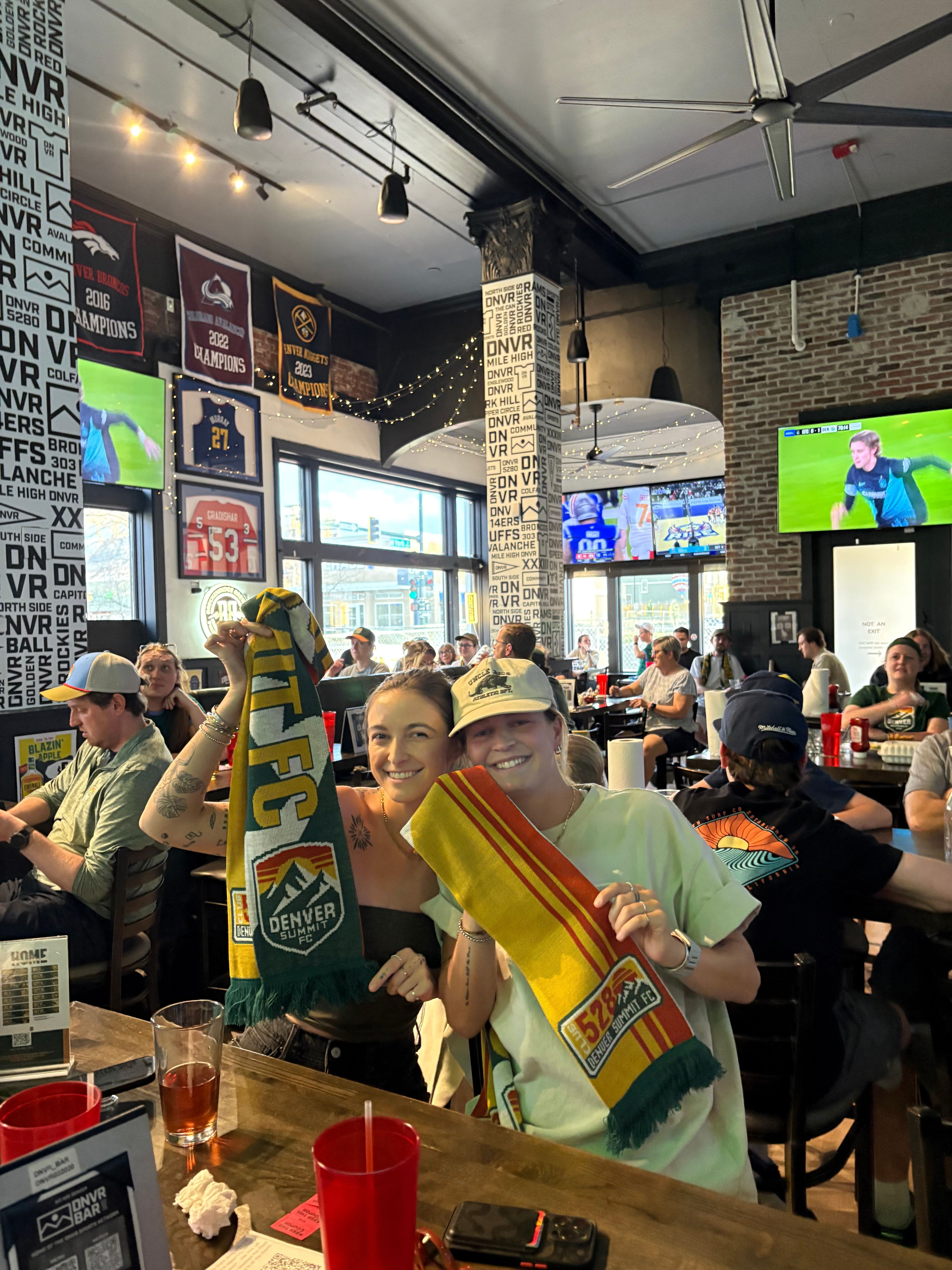 People gather in a sports bar, watching a game on multiple screens, some holding drinks and snacks, with a brick wall background.