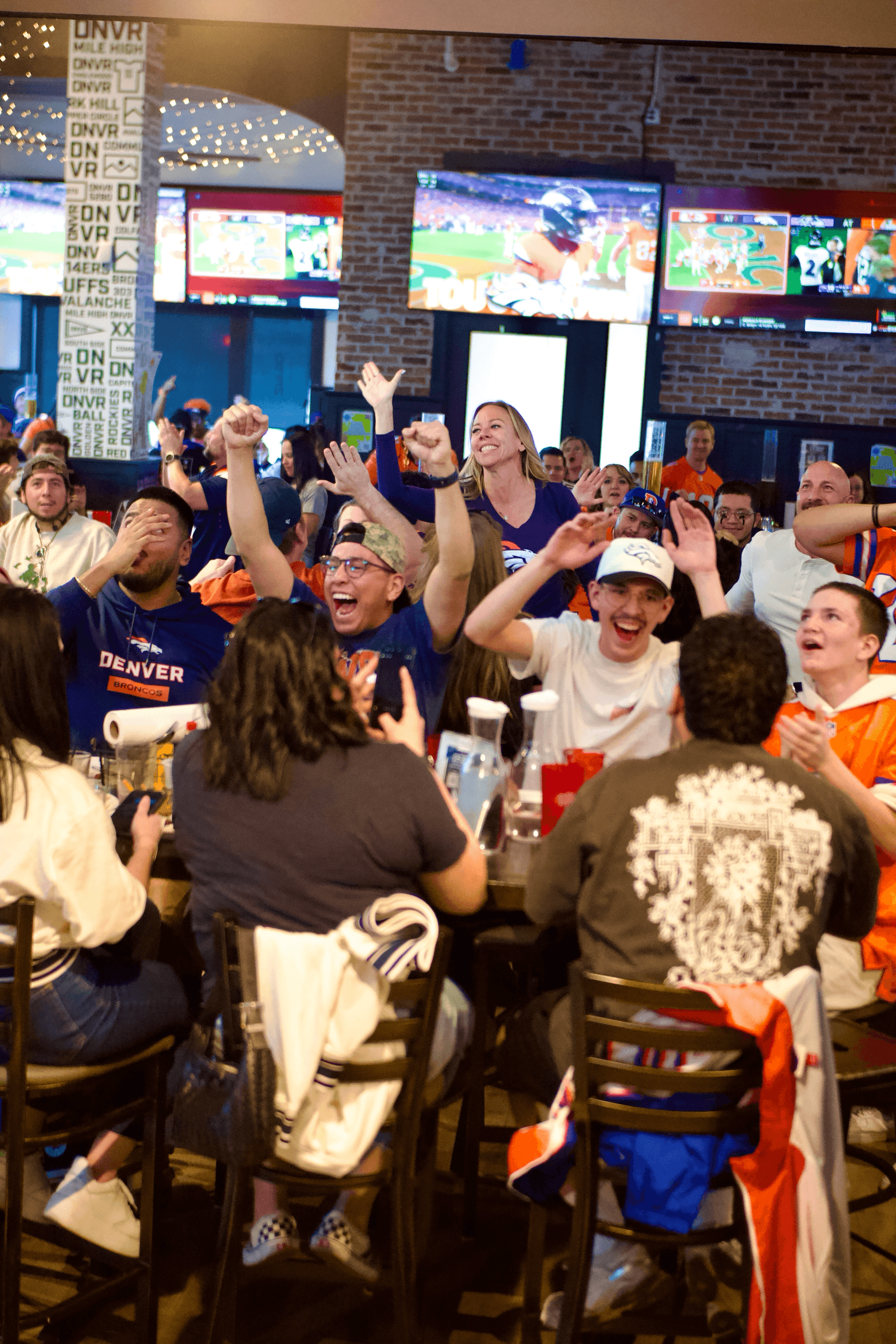 Group of people celebrating and cheering at a social event, some wearing Denver shirts, in a lively indoor setting.
