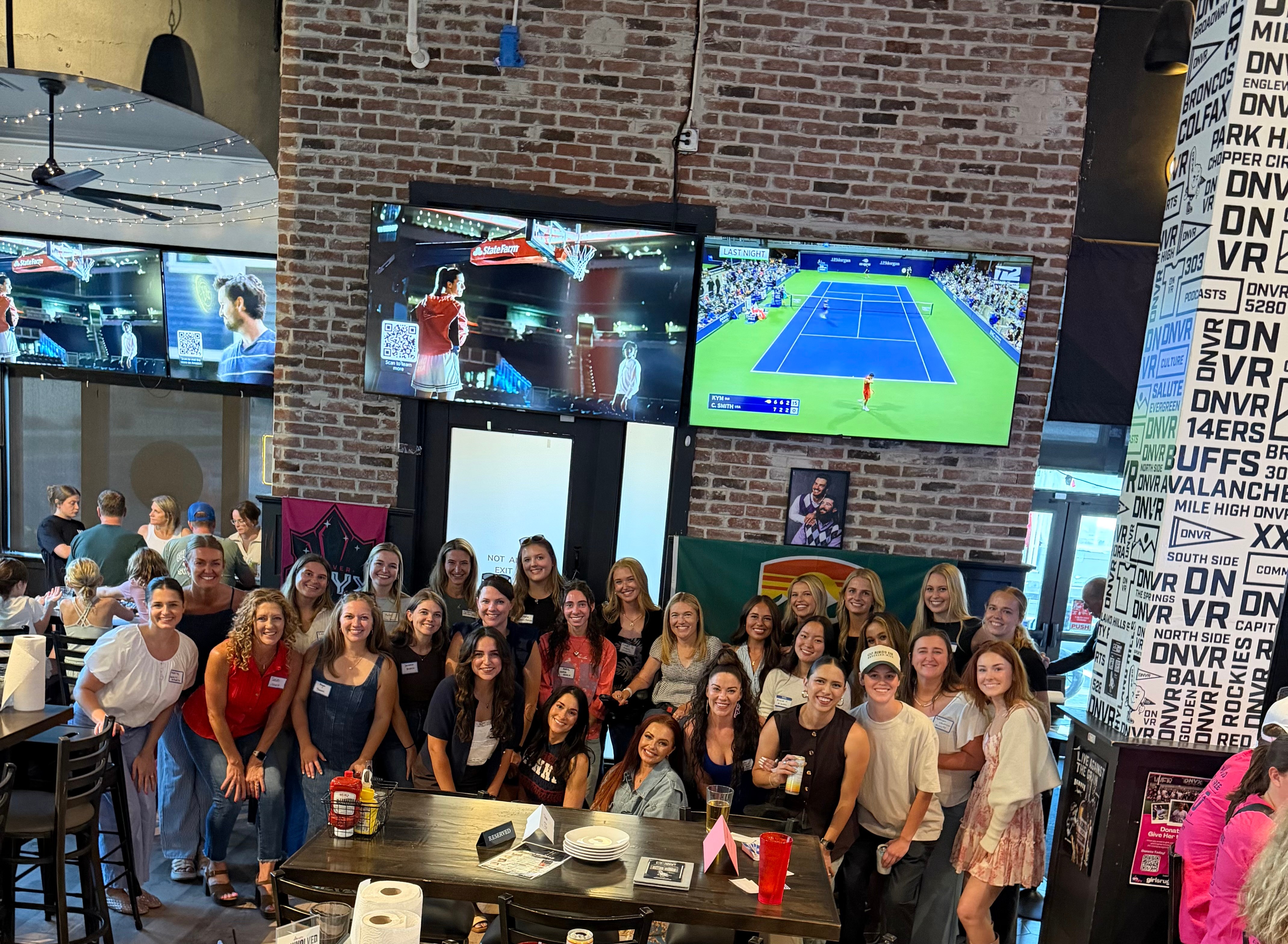 Group of people gathered in a room with brick walls, two large TV screens showing tennis matches, and a table with food and drinks.