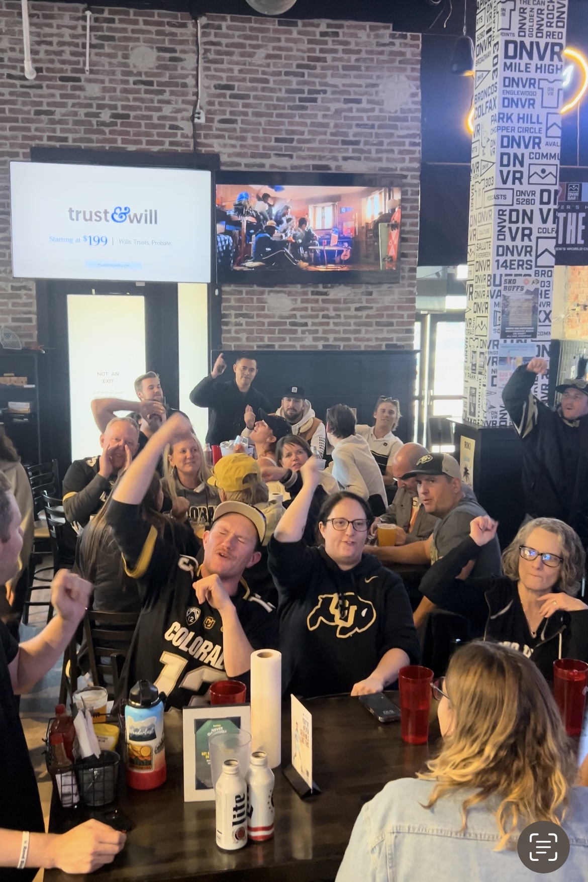 People gathered in a bar or restaurant, some raising their fists, with brick walls and screens in the background.