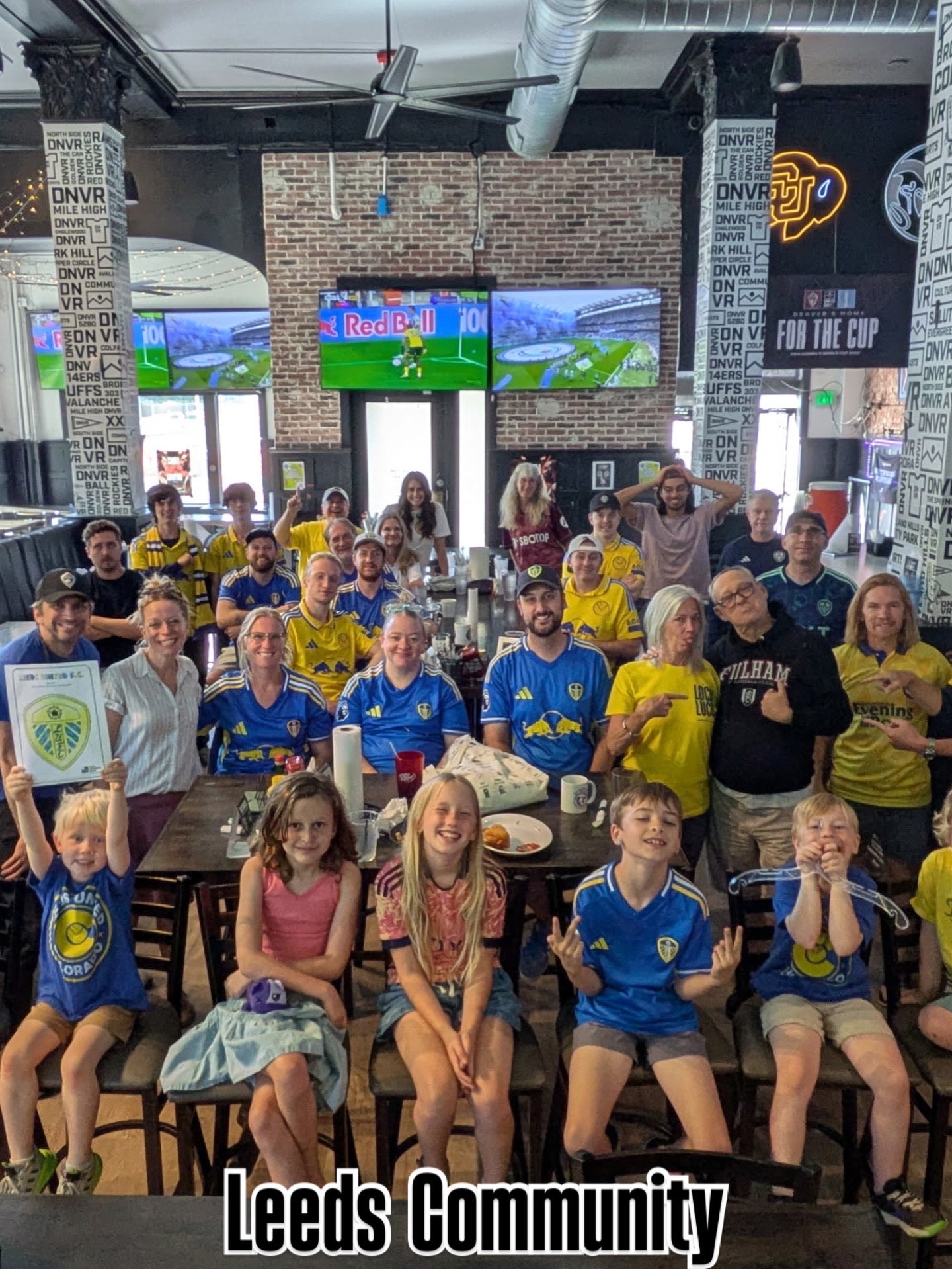 Group of children and adults gathered indoors, some wearing Leeds Community shirts, with screens and banners in the background.