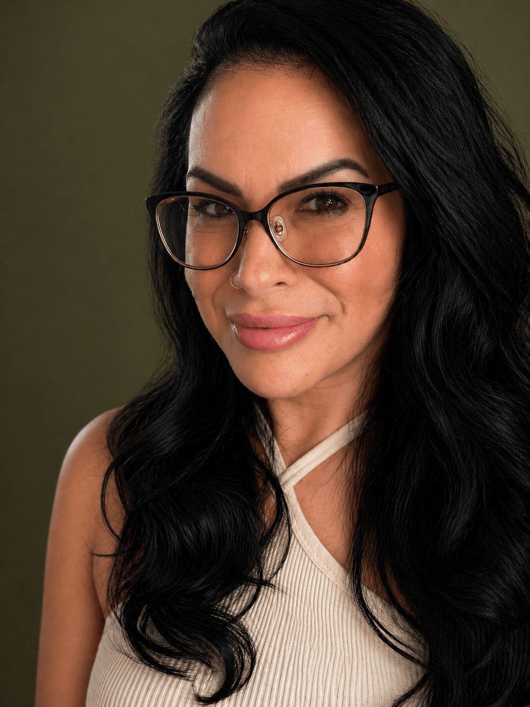 Woman with long dark hair, glasses, and a light-colored top, smiling softly against a dark background.