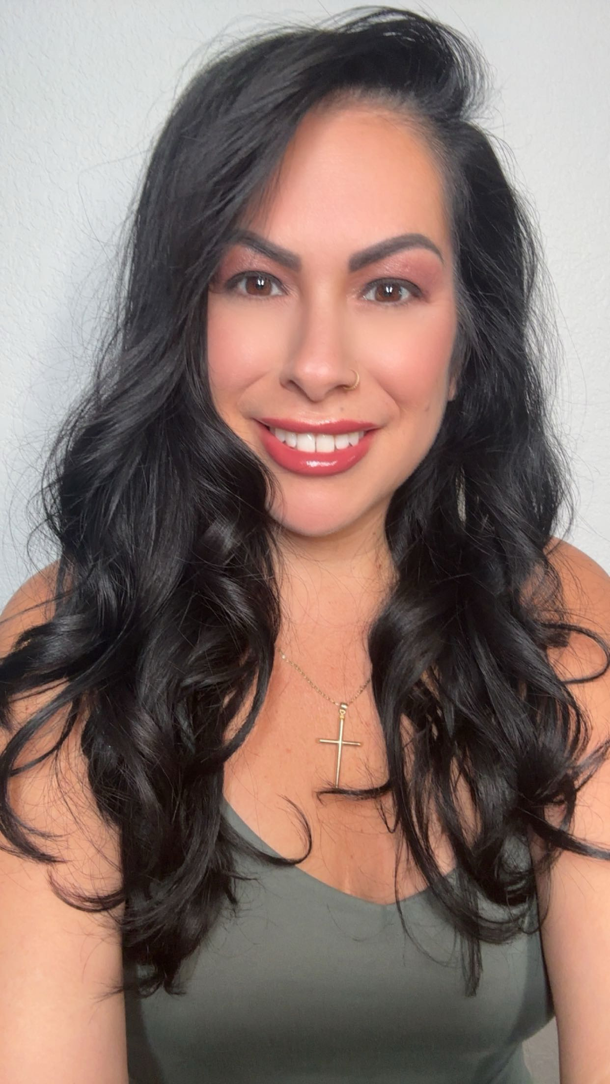 Woman with long dark wavy hair smiling, wearing a gray top and a cross necklace, against a plain background.