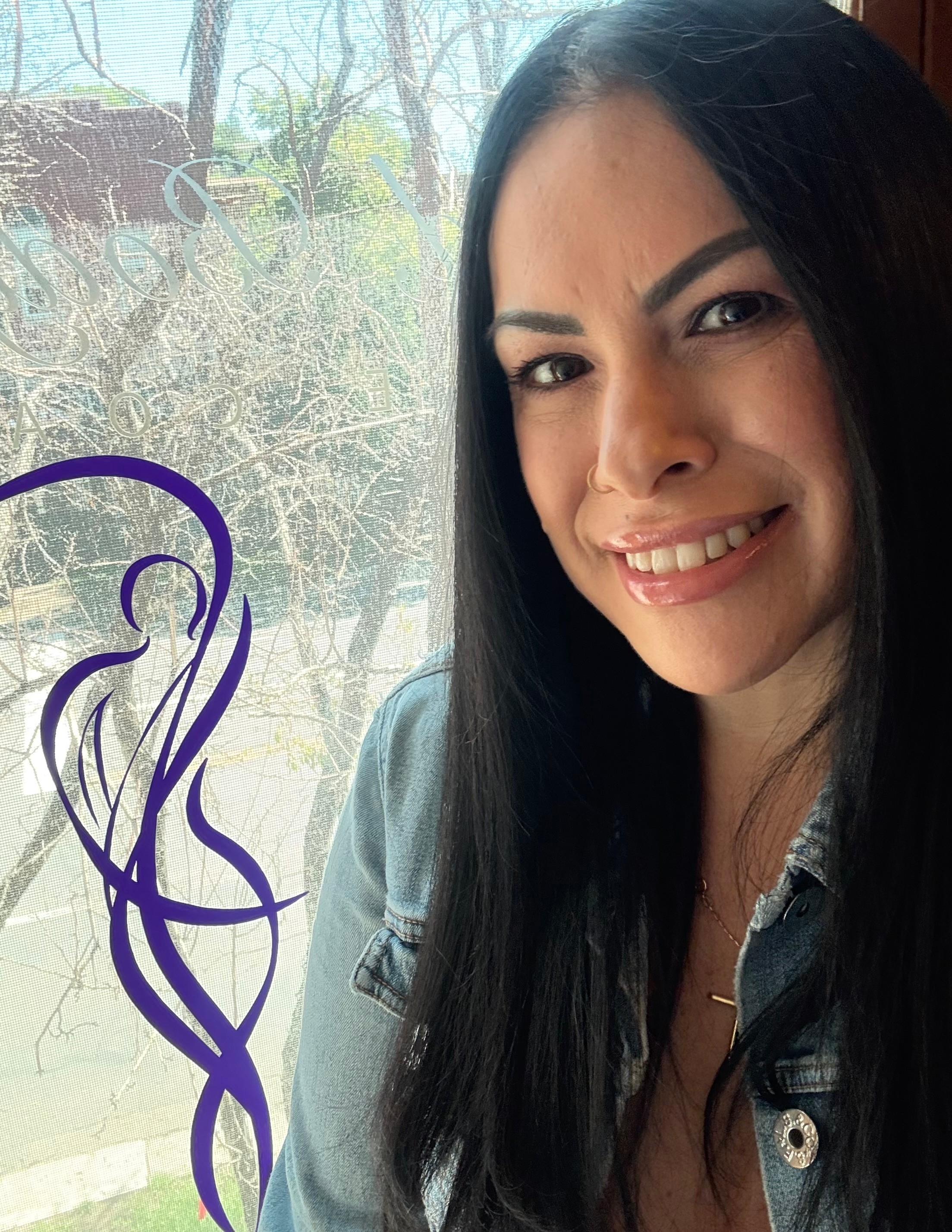 Young woman with long dark hair smiling near a window with trees outside.