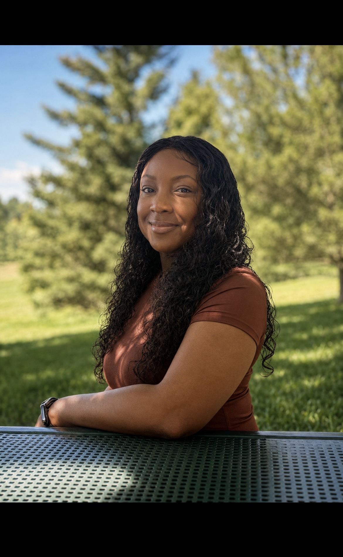 Young woman with long curly hair smiling outdoors, sitting at a table with trees and grass in background.