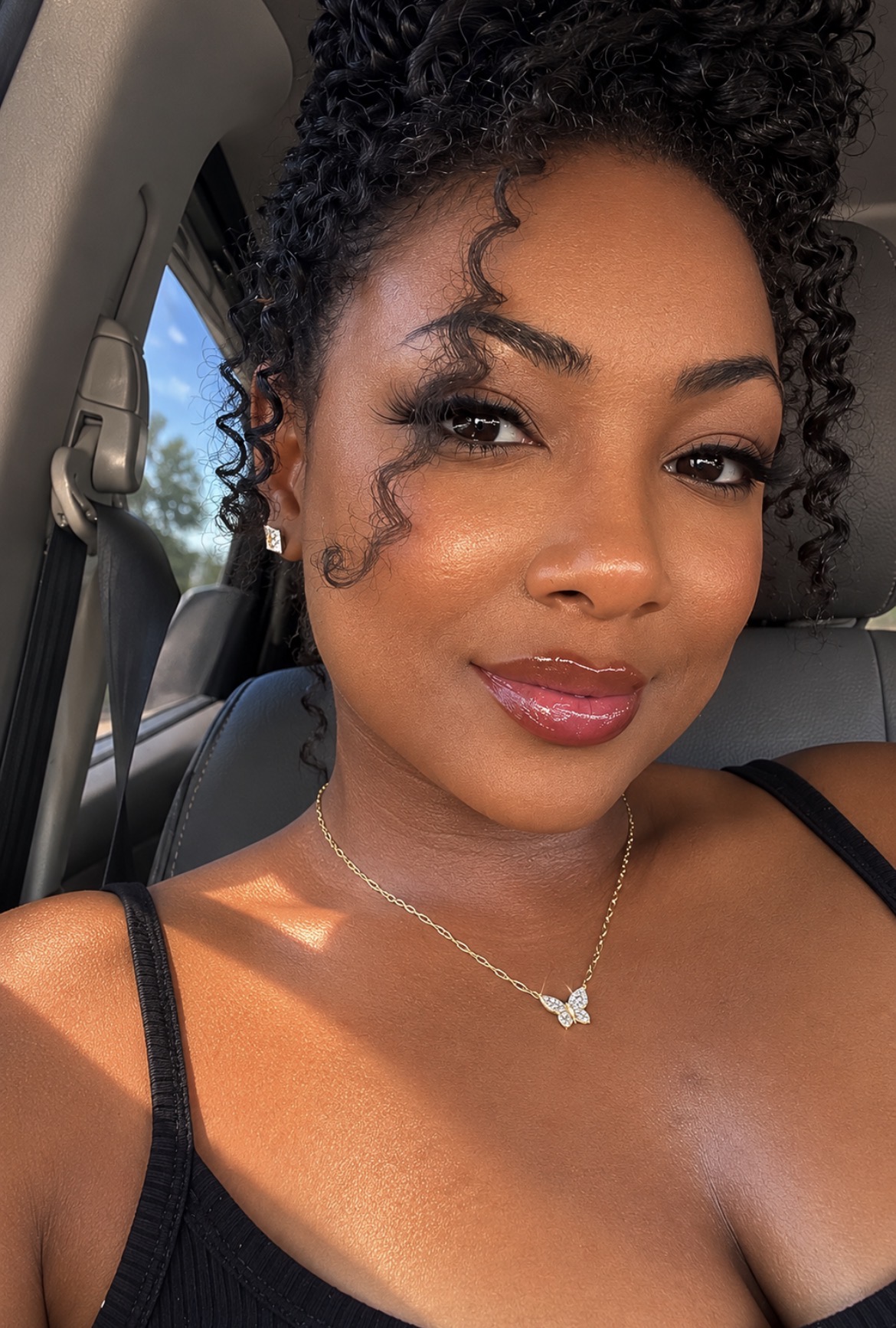 Close-up of a woman with curly hair, wearing a black top and butterfly necklace, smiling inside a car.
