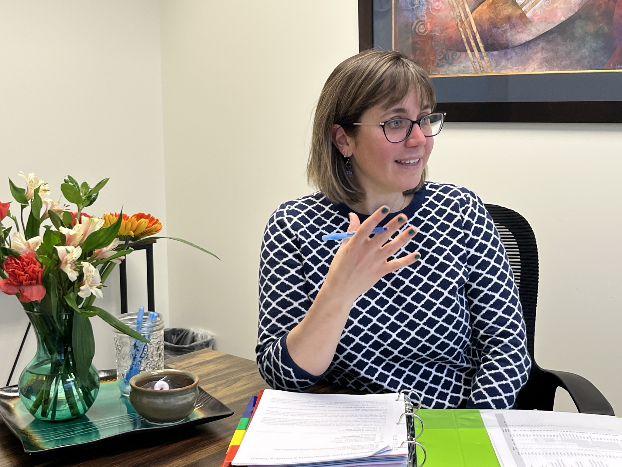 Woman with glasses and short hair, wearing a patterned top, sitting at a desk with papers, flowers, and a painting in the background.