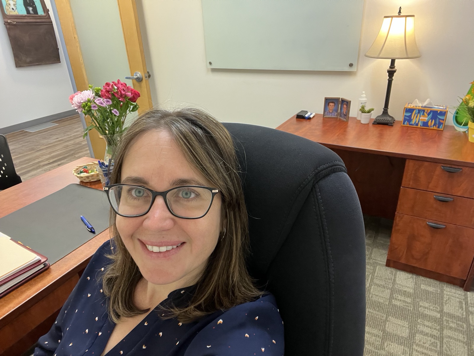 Woman with glasses smiling in an office, desk with flowers, lamp, and framed photos in background.