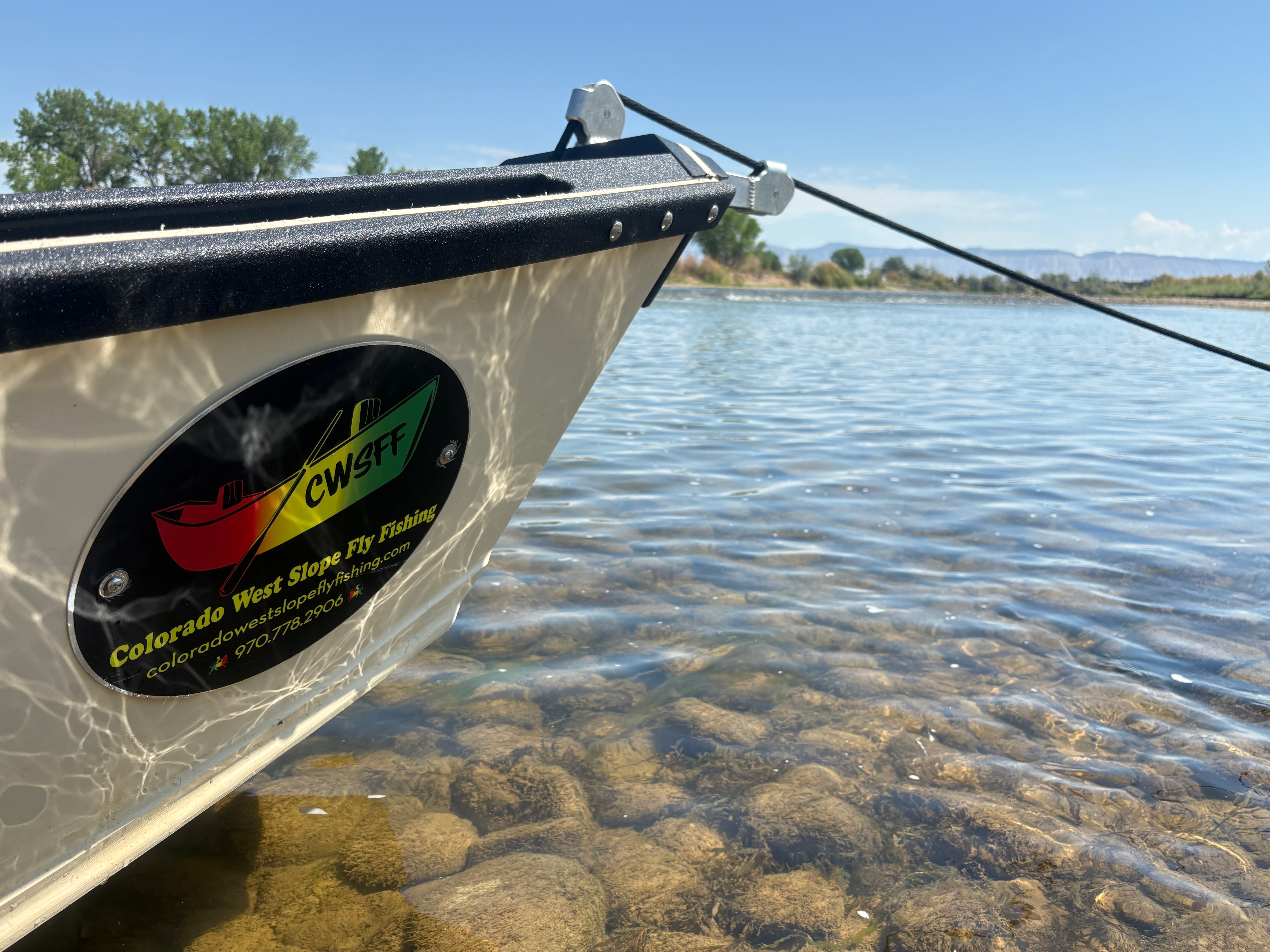 Close-up of a boat's side with a logo, floating on clear water with rocks visible underneath, and a distant landscape.