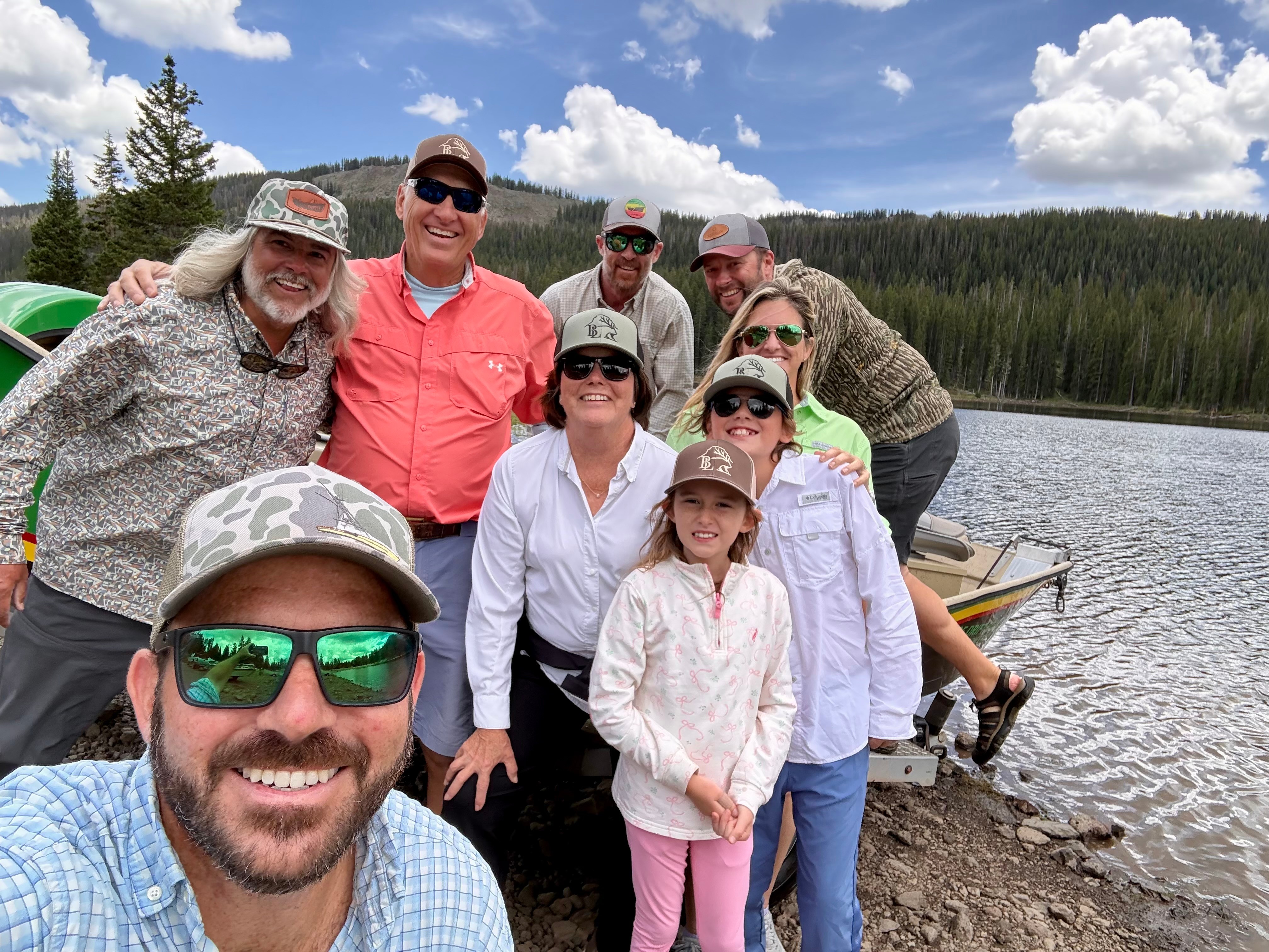 Group of nine people outdoors near a lake with trees and mountains in the background, smiling and posing for a photo.
