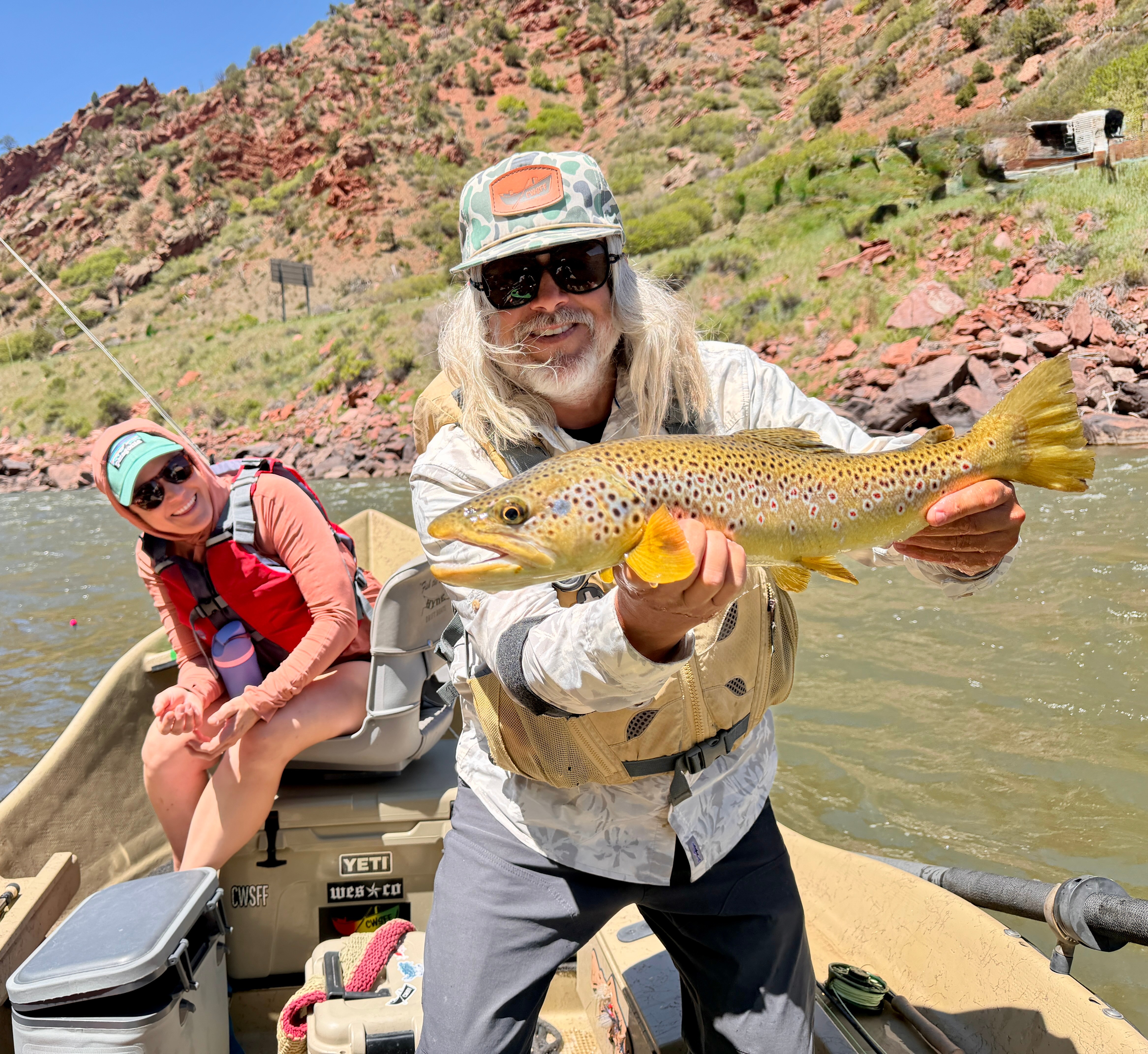 Man holding a fish on a boat with a woman sitting nearby, river and hillside in background.