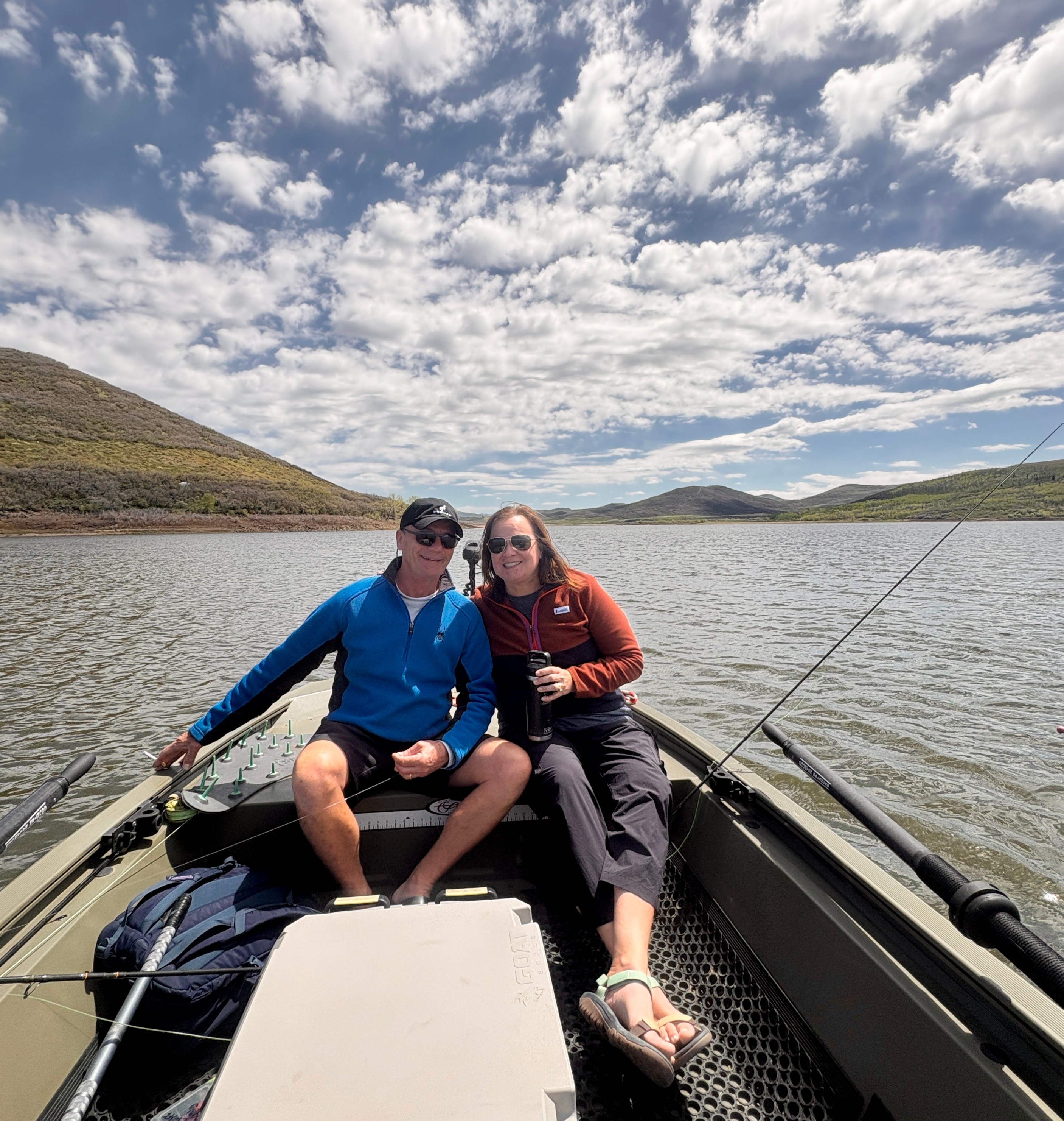 Two people sitting in a boat on a lake with hills and a partly cloudy sky in the background.