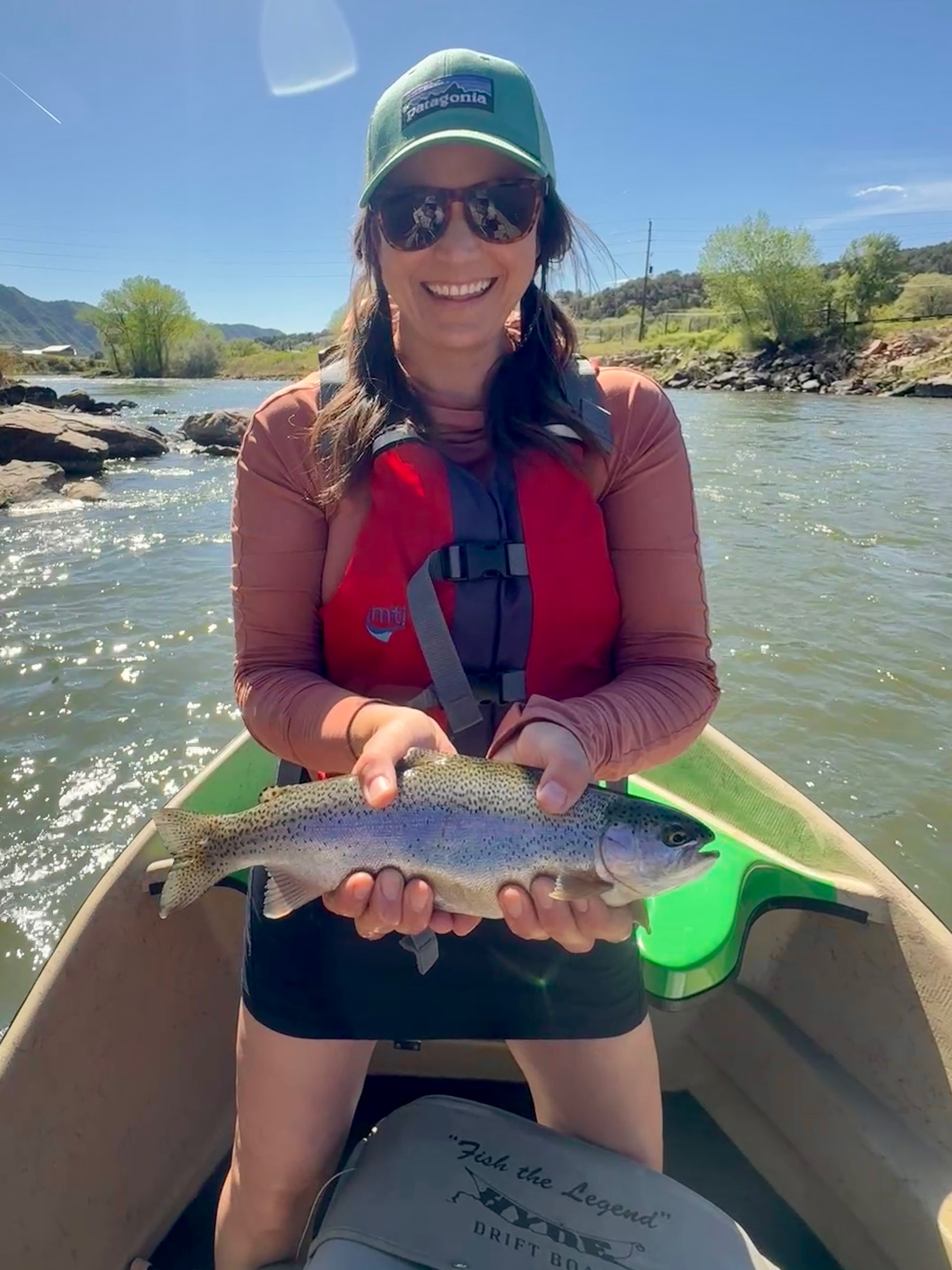 Woman in sunglasses and hat holding a fish in a boat on a river, smiling, with trees and blue sky in background.
