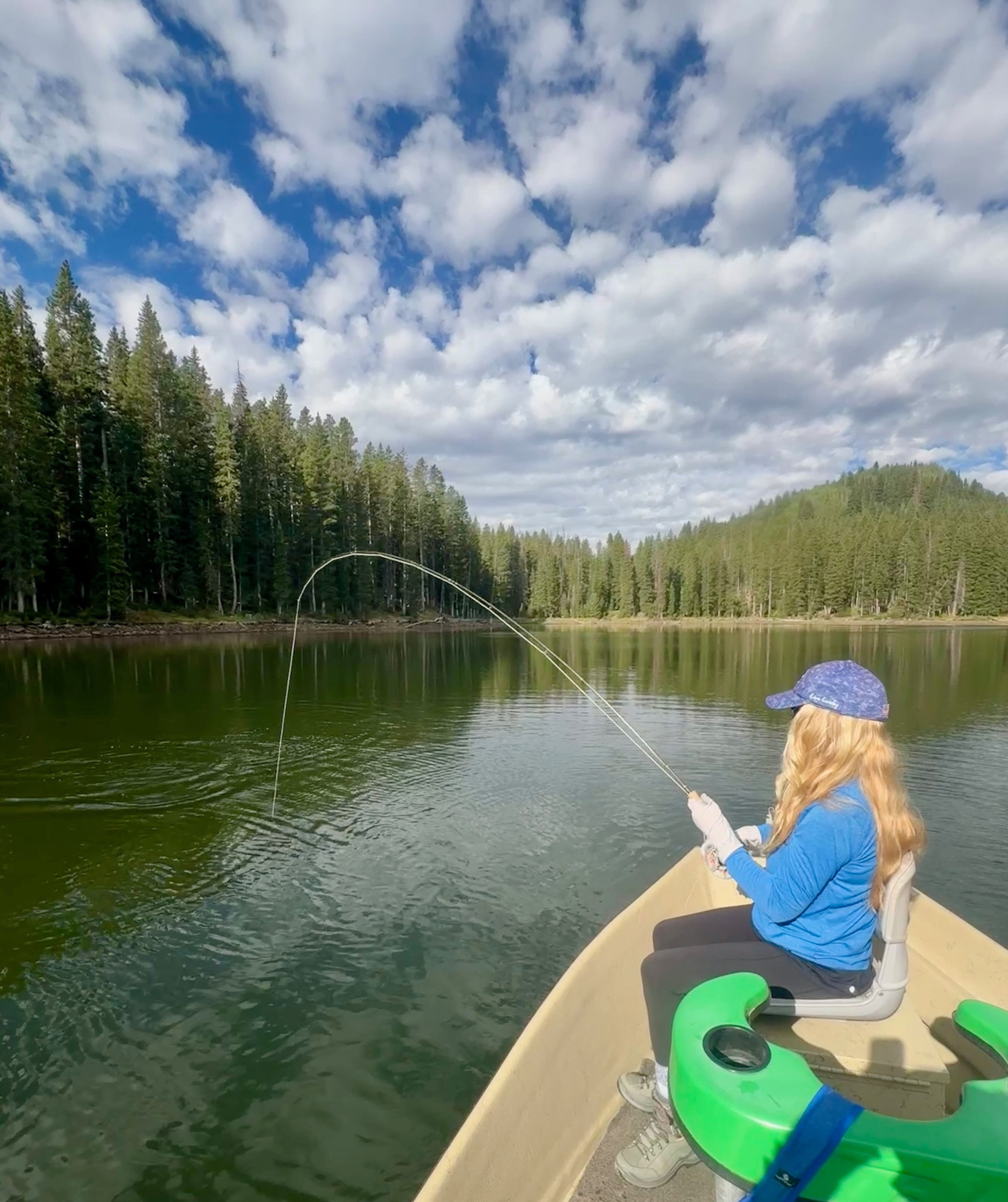 Person with long hair wearing a blue jacket and cap sitting in a boat, fishing in a lake surrounded by trees under a partly cloudy sky.