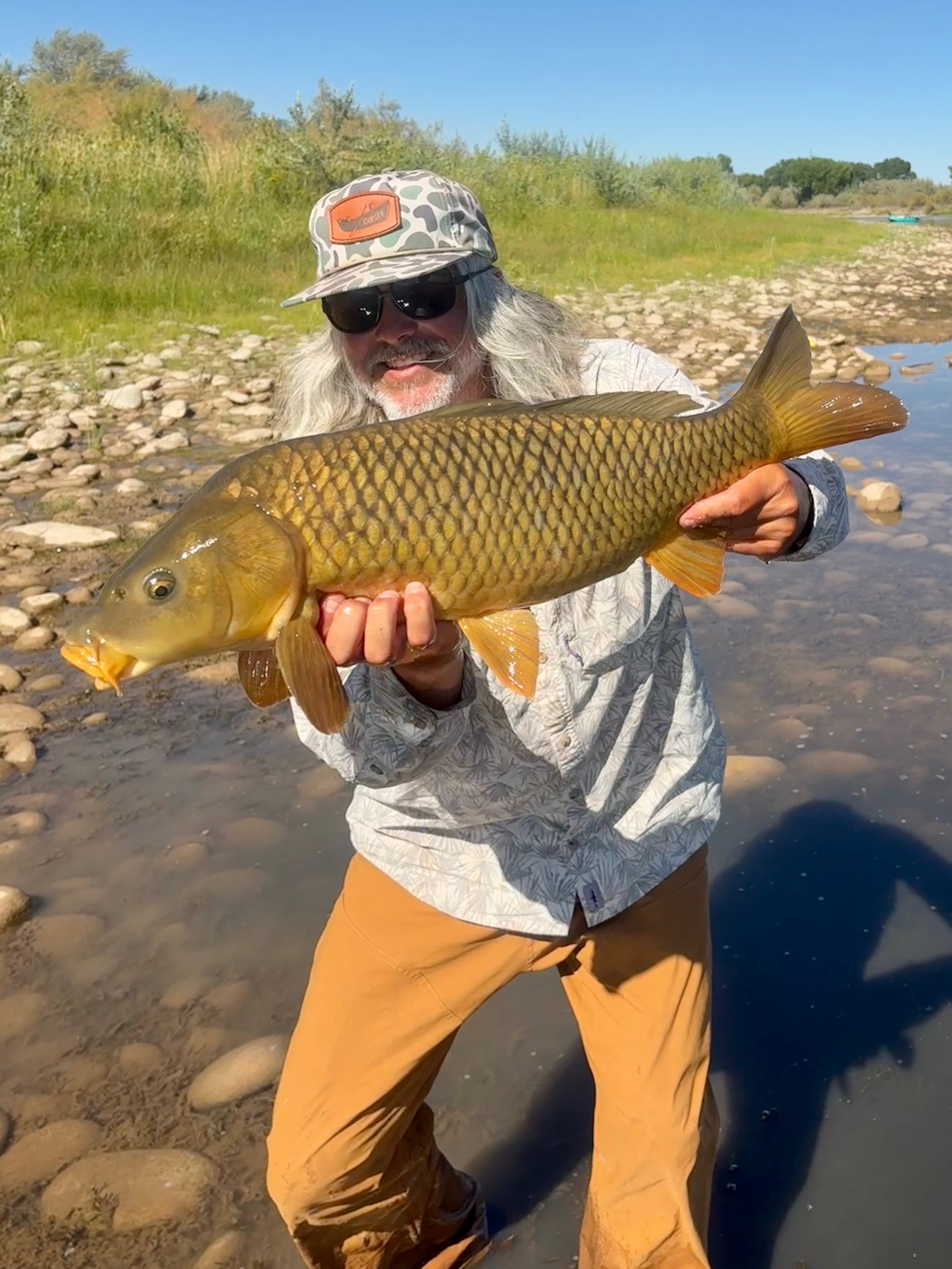 Person holding a large fish outdoors on a rocky riverbank, wearing a hat and sunglasses, with greenery and blue sky in background.