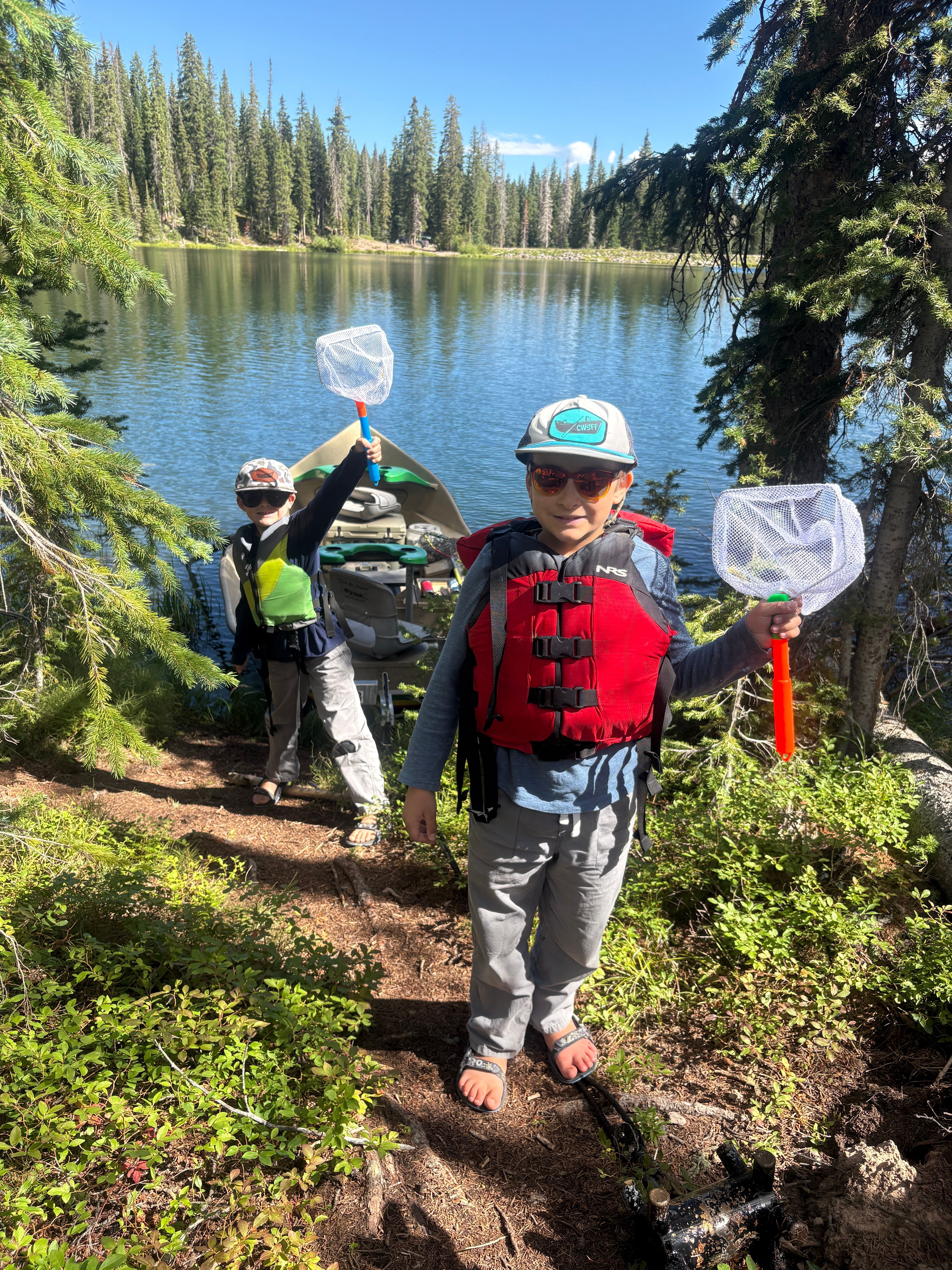 Two children with backpacks and nets near a lake, surrounded by trees and greenery, on a sunny day.