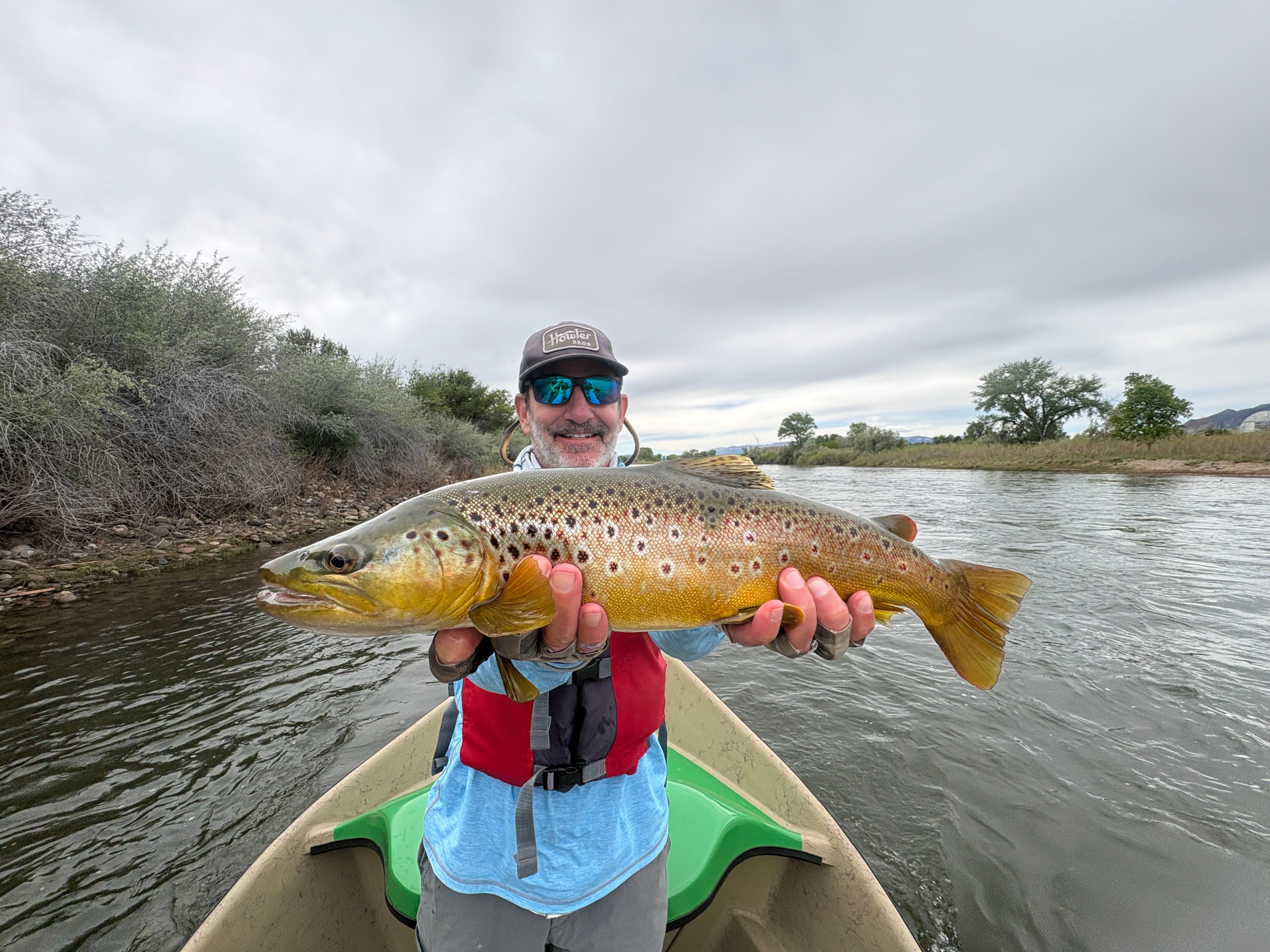 Person holding a large brown and yellow fish on a boat in a river, with cloudy sky and trees in background.