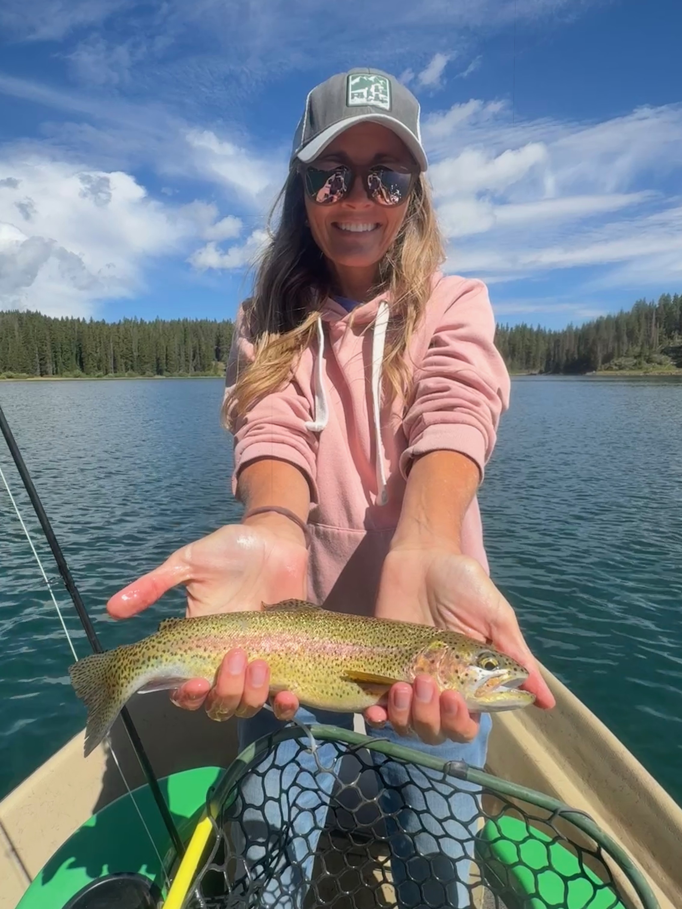 Woman in pink hoodie and sunglasses holding a fish on a boat, lake, and trees in background.