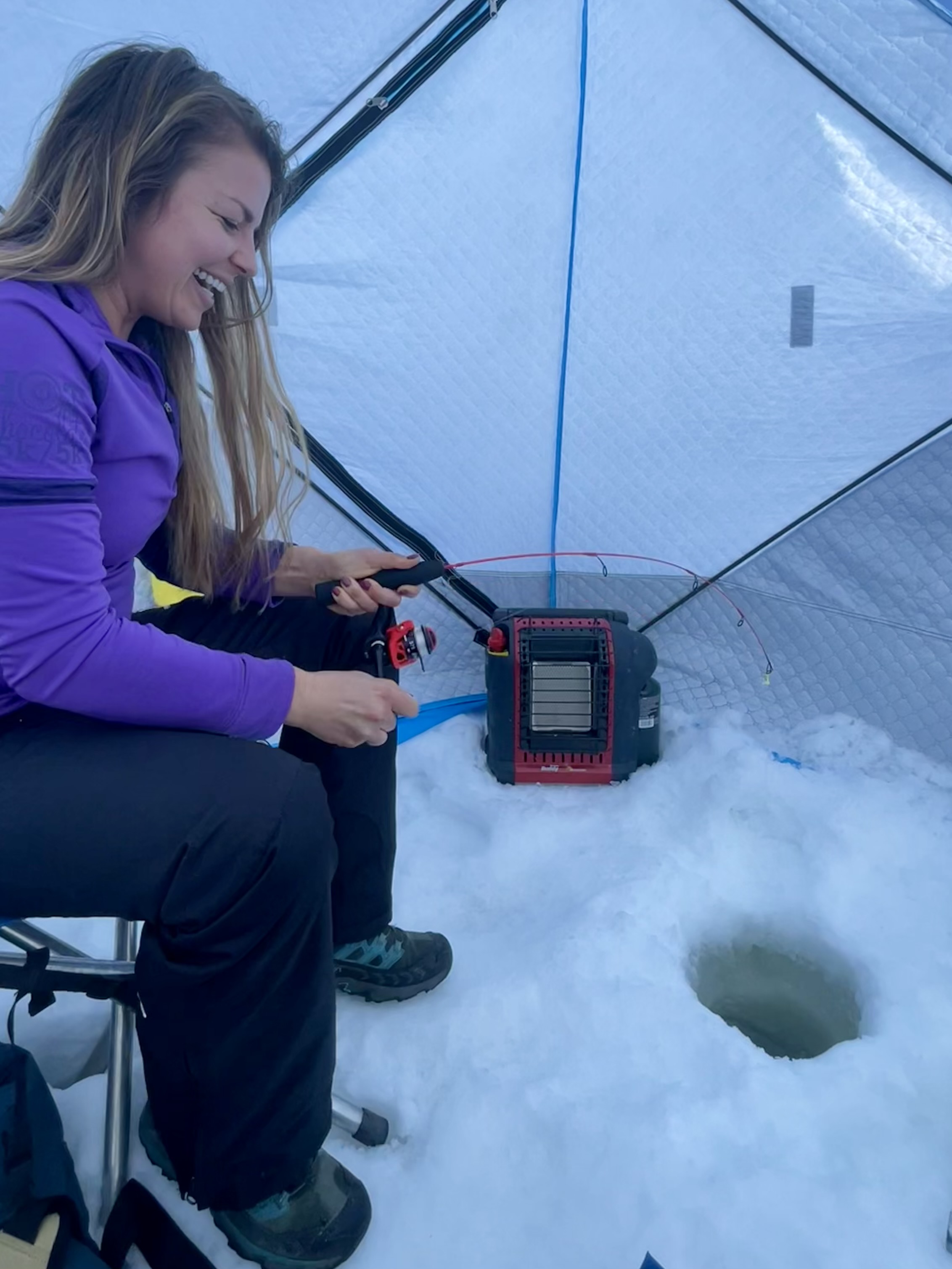 Woman sitting inside a tent, ice fishing with a portable heater and fishing hole in snow, smiling.