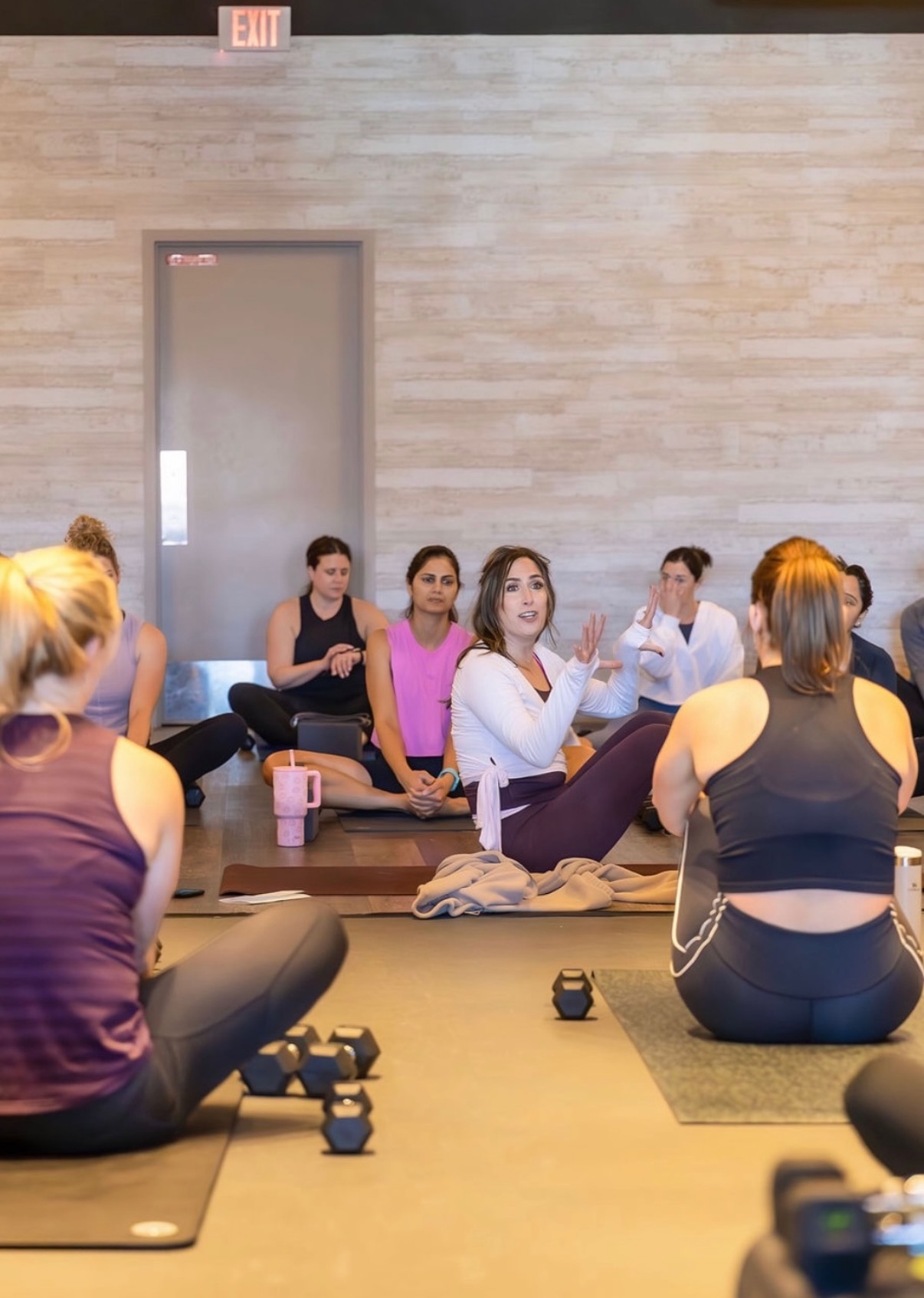 Group of women participating in a yoga or fitness class indoors, sitting on mats with fitness equipment nearby.