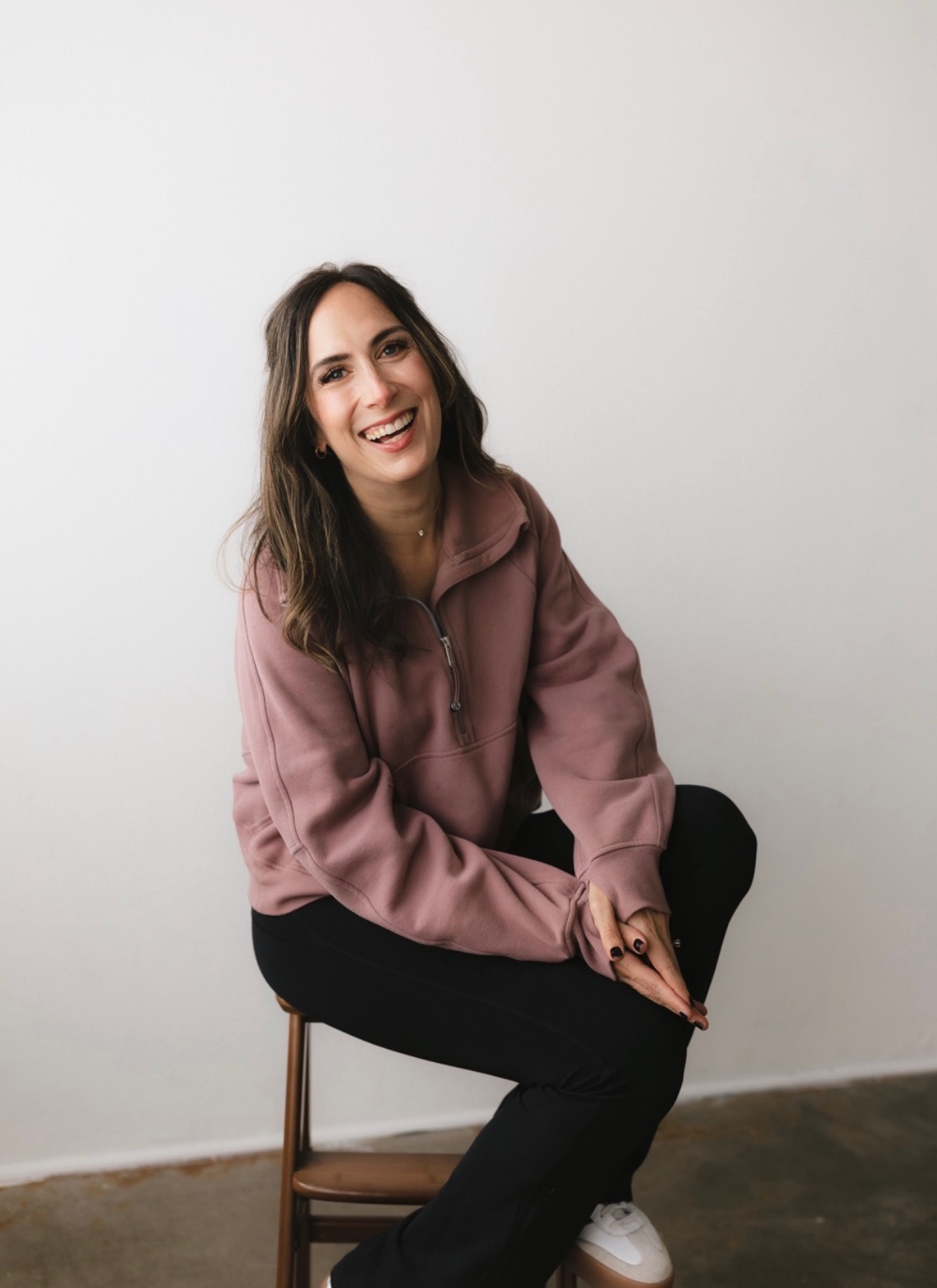 Young woman with long wavy hair smiling, sitting on a wooden chair against a plain white wall, wearing a pink hoodie and black pants.