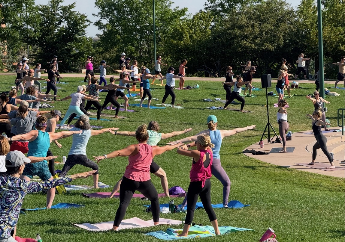 Group of people practicing yoga outdoors on mats in a park with trees and a paved area, some with arms extended.