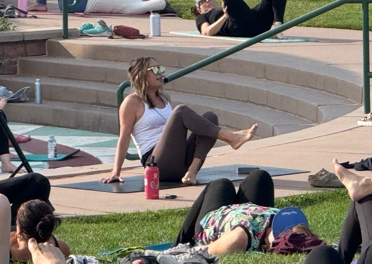 Woman sitting on steps outdoors, wearing sunglasses, white tank top, and brown pants, with a pink water bottle nearby.