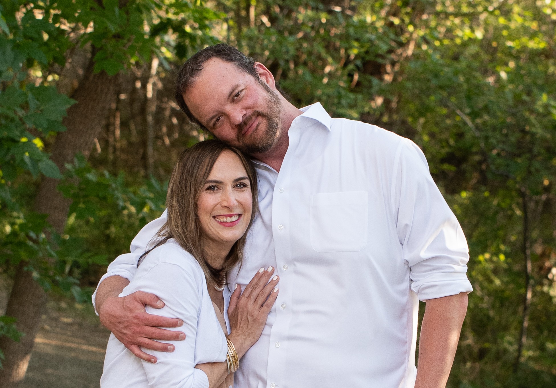 A smiling couple hugging outdoors with trees in the background.