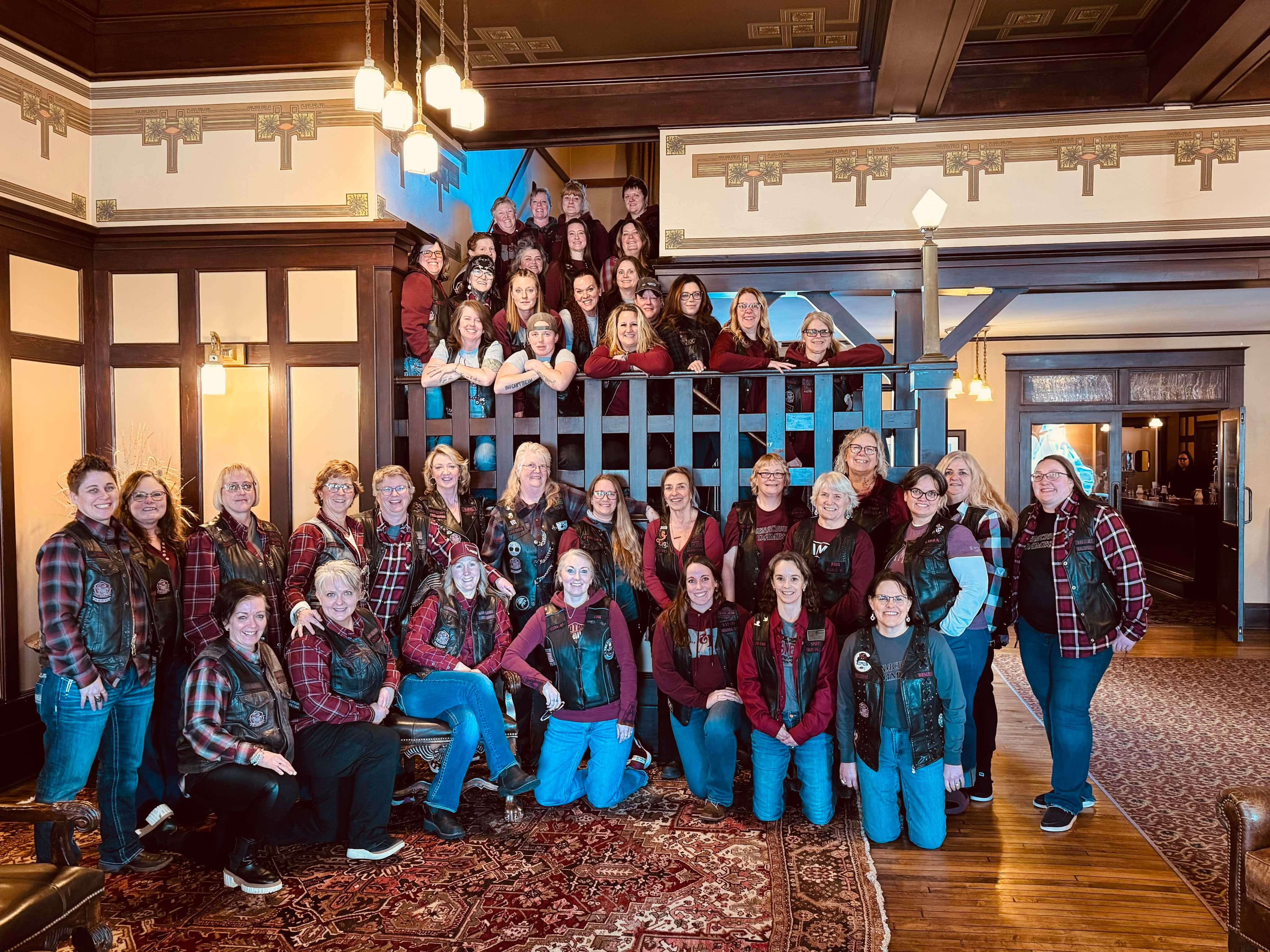 Group of people gathered in a room with wooden paneling and a patterned carpet, some standing on a staircase, smiling.