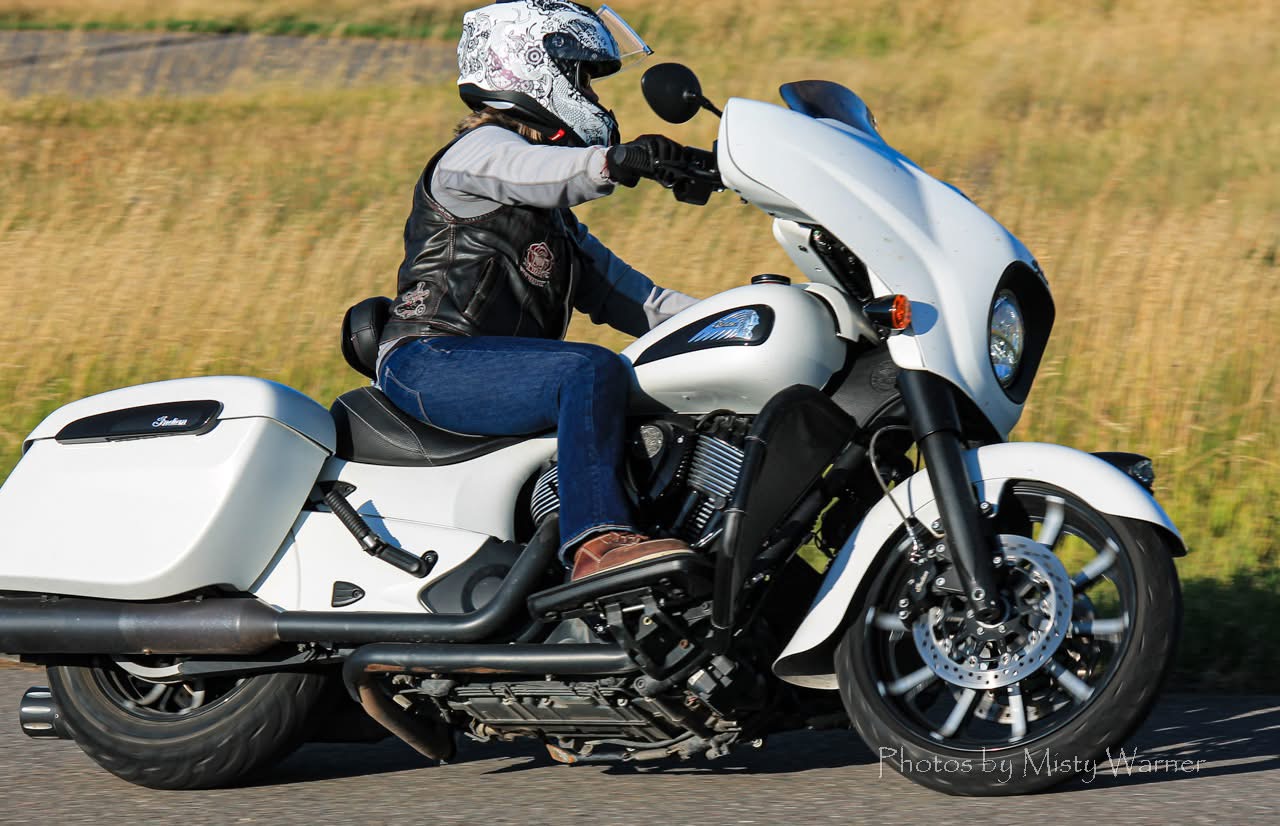 Person riding a white motorcycle with a helmet, black jacket, and jeans on a road with grassy background.
