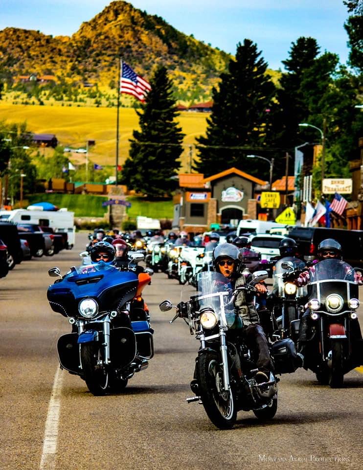 Motorcycles and riders in a parking lot with trees, buildings, and a mountain in the background.