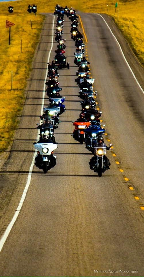 A long line of motorcycles riding on a two-lane road through a grassy landscape, with some riders wearing helmets and gear.