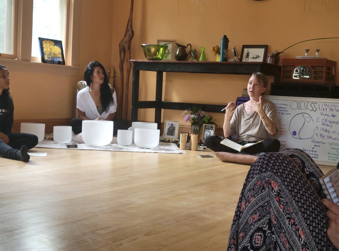 People sitting around a table in a room with a woman speaking near a whiteboard, with various objects on a shelf behind her.