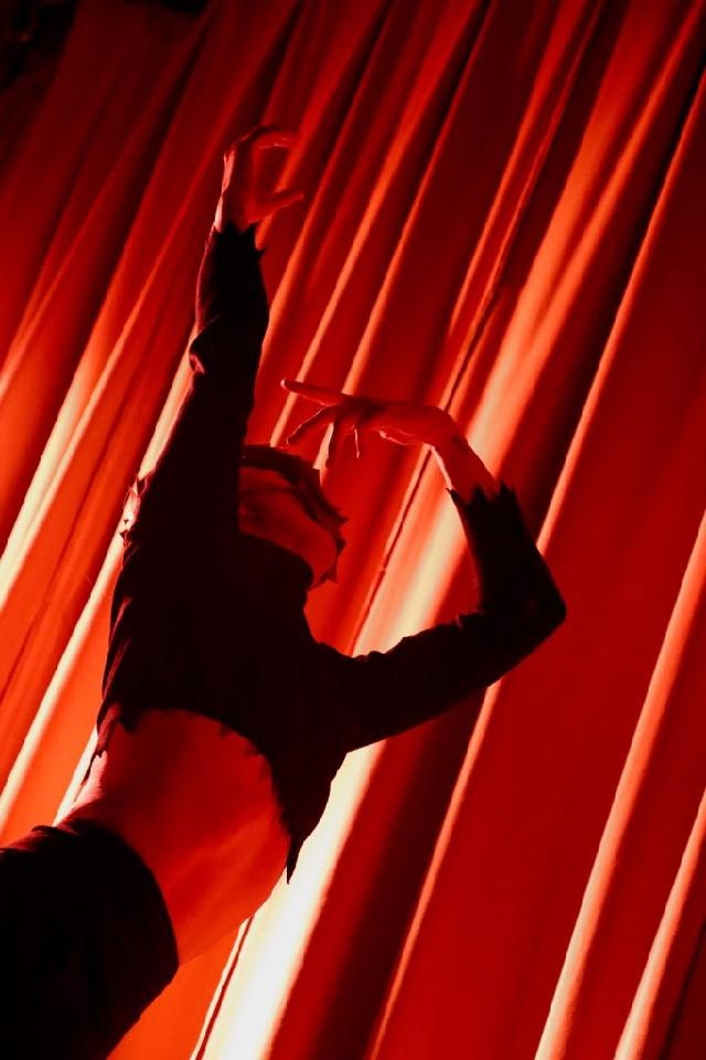 Person performing a dance move with arms raised and curved, against red stage curtains.
