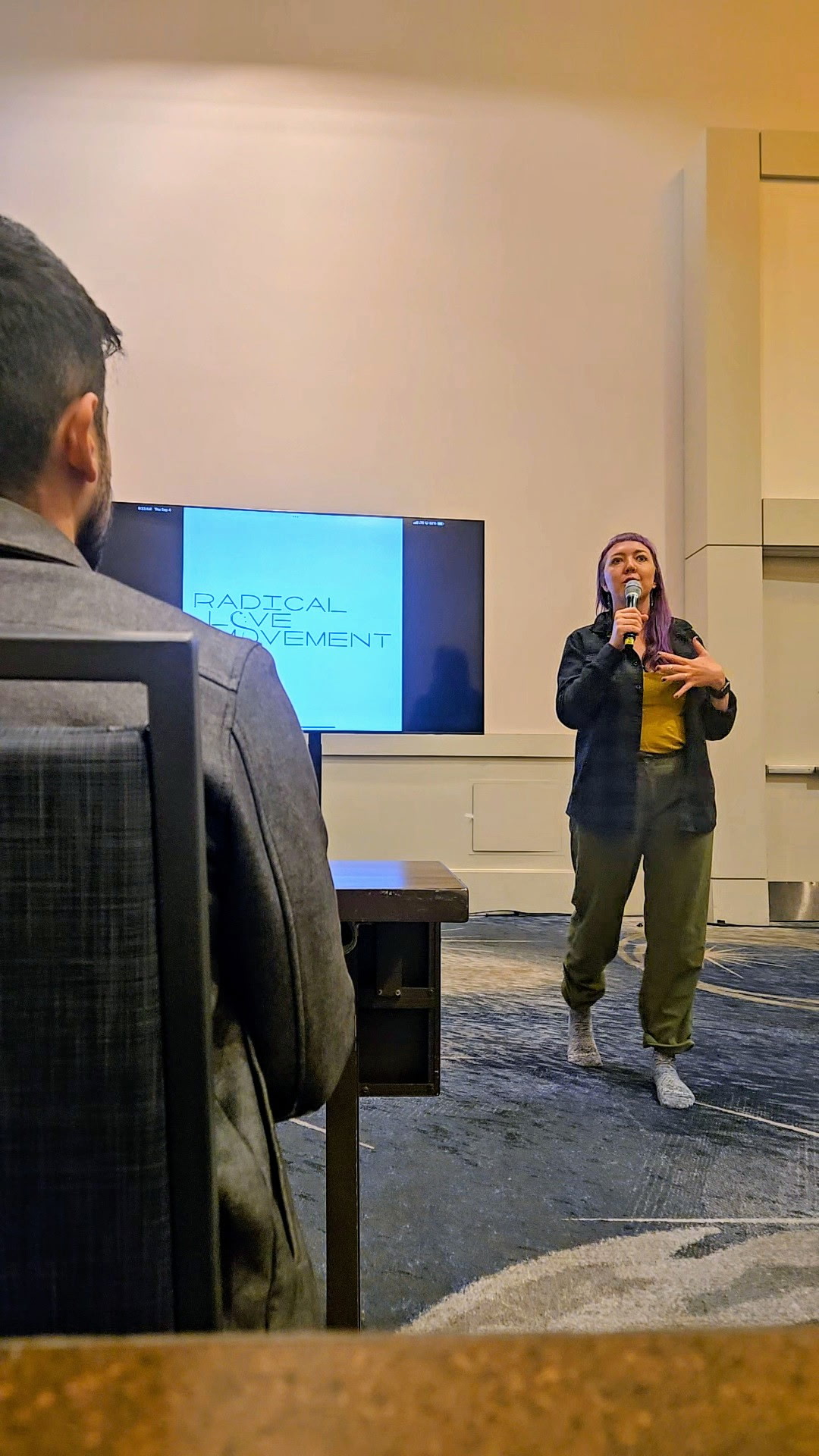 Person standing and speaking to an audience in a conference room with a presentation screen behind.
