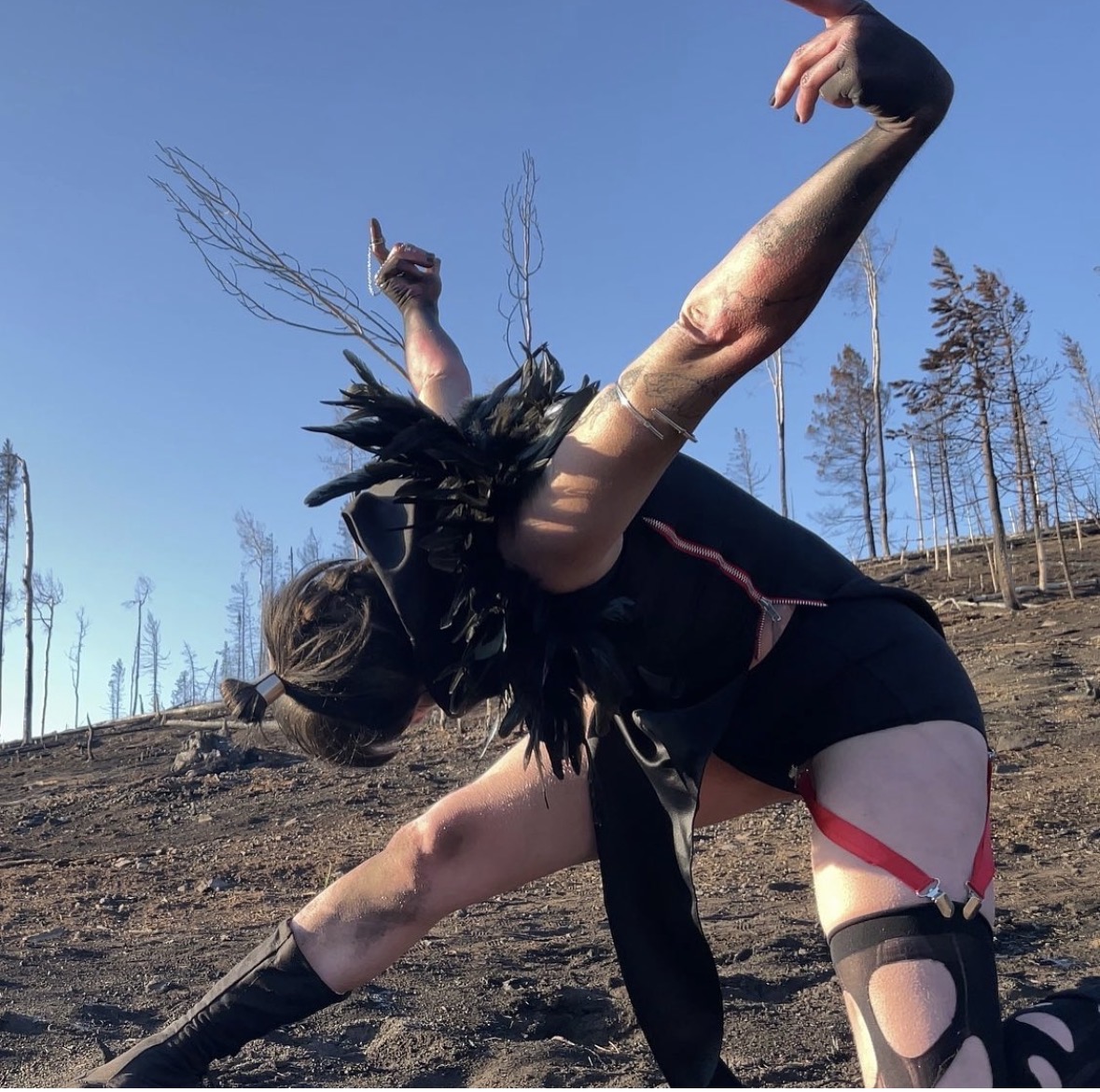 Person performing a handstand outdoors on a dirt surface with trees in the background, wearing black clothing and accessories.