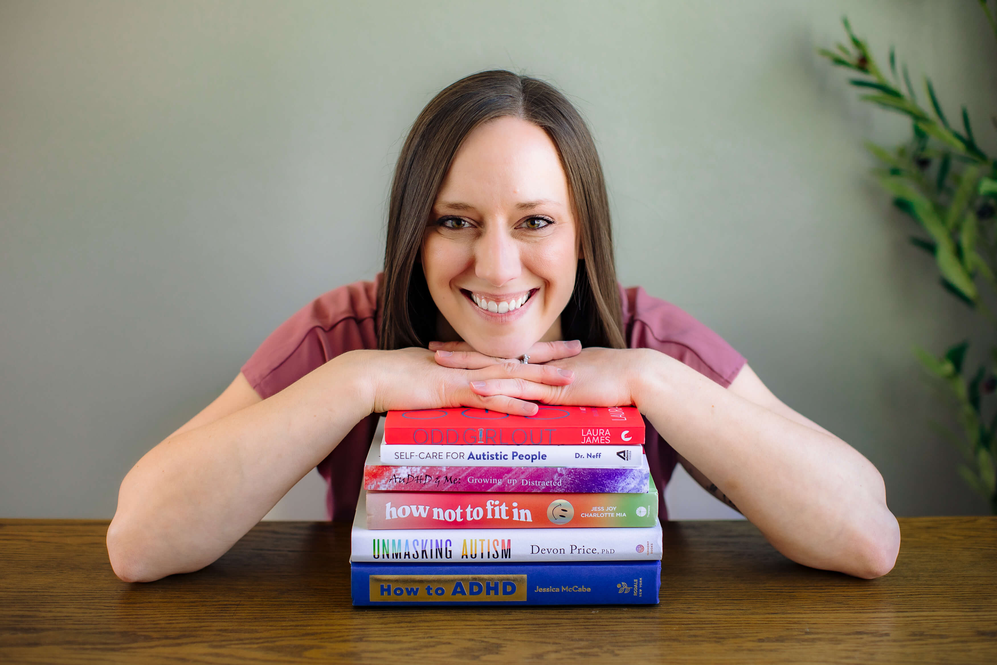 Smiling woman resting her chin on stacked colorful books on a table, with a green plant in the background.