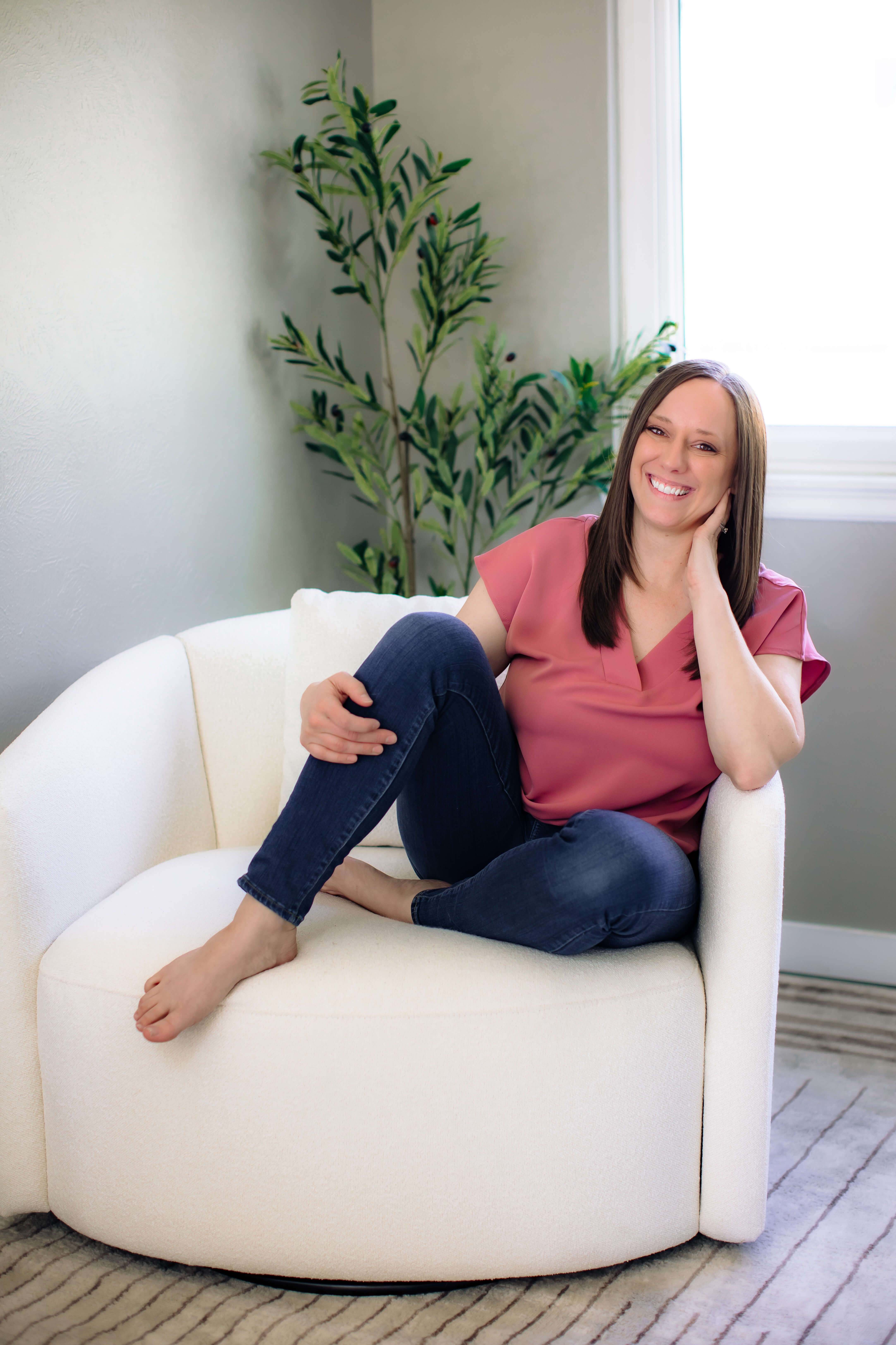 Woman sitting on a white sofa, smiling, with a large plant and window behind her.