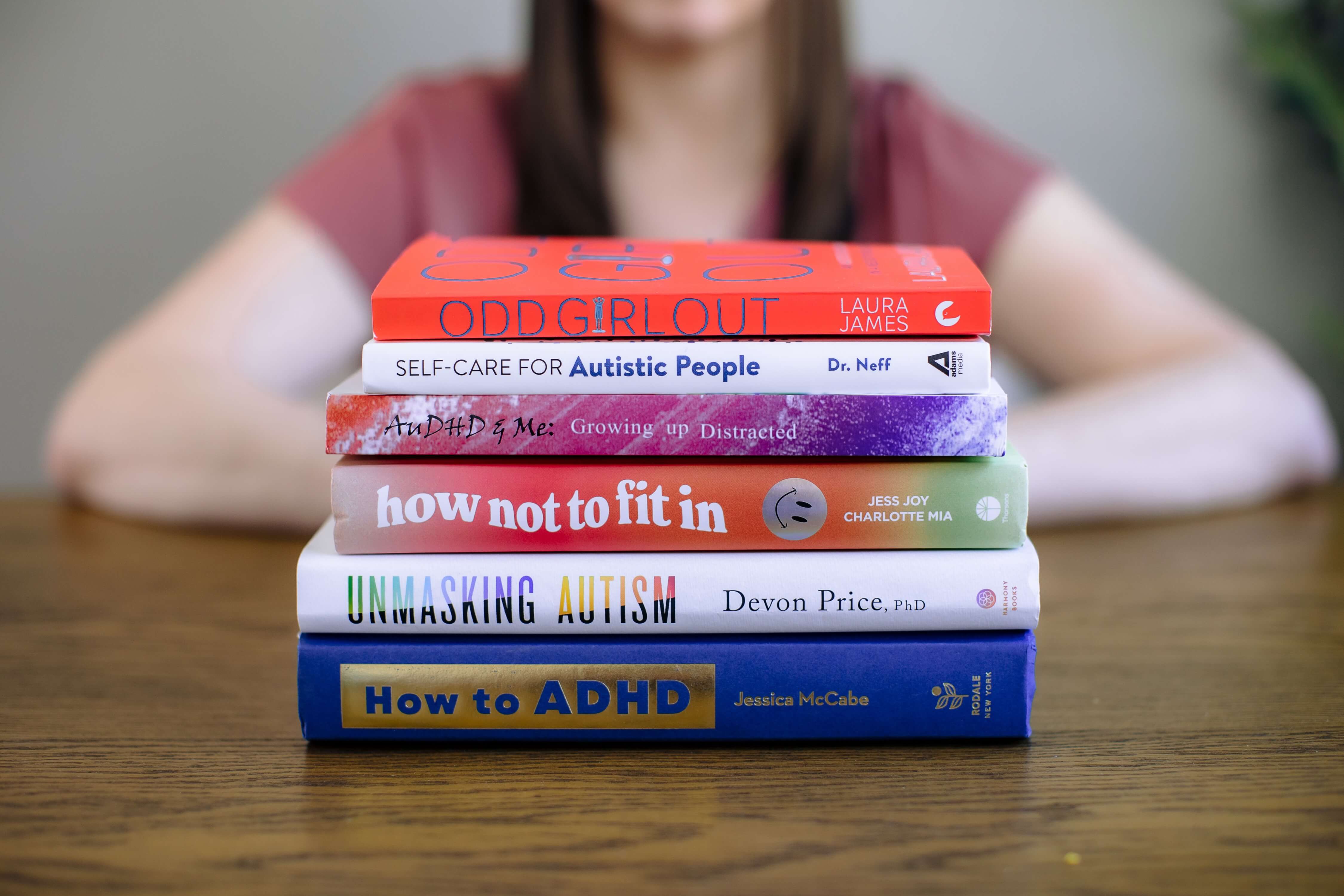 Stack of five books on a table with a person blurred in the background, arms resting on the table.