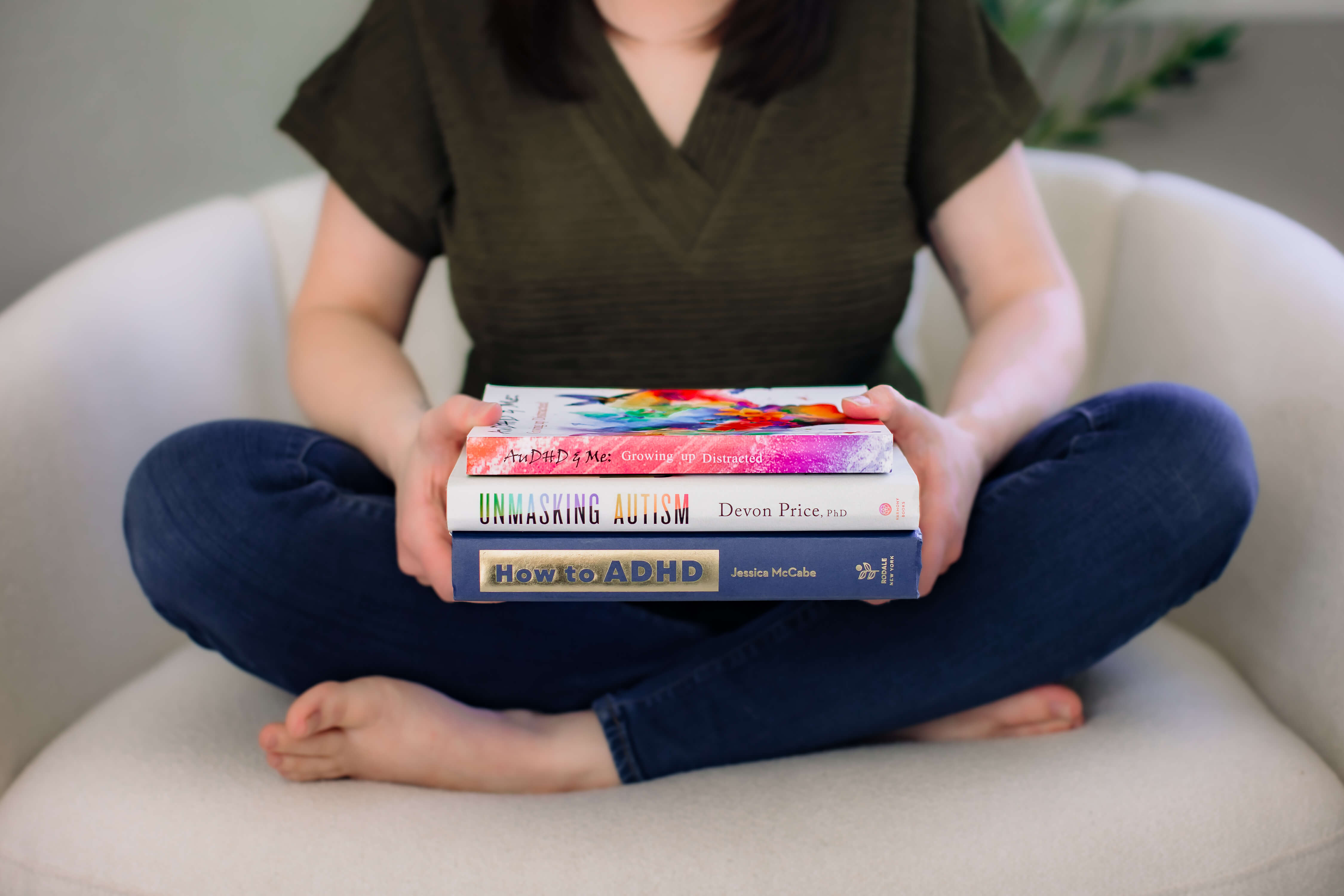 Person sitting cross-legged on a sofa holding a stack of books, with only torso and legs visible.